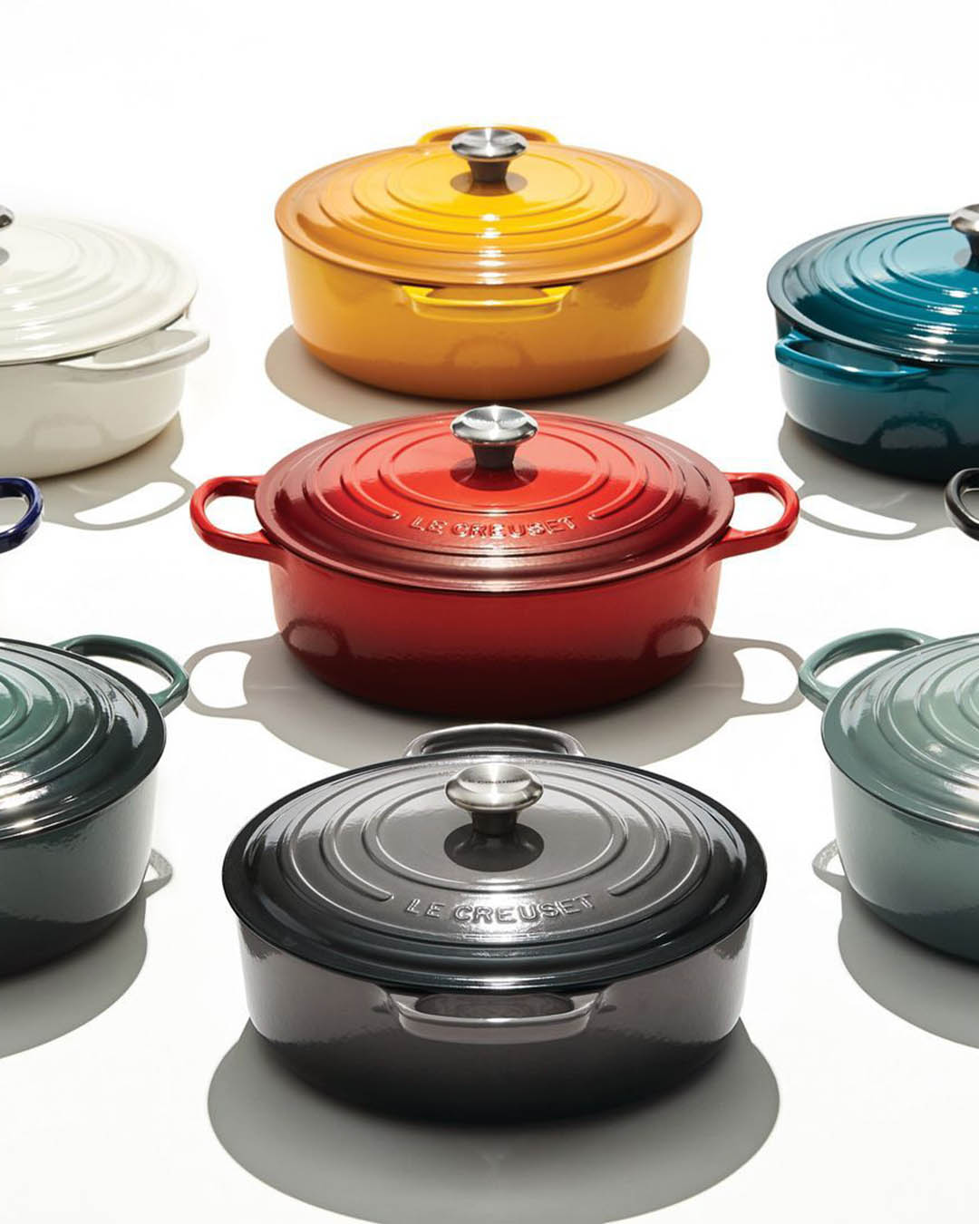 Top-down view of colourful Le Creuset cast-iron casserole dishes (red, yellow, teal, grey) arranged on a white background