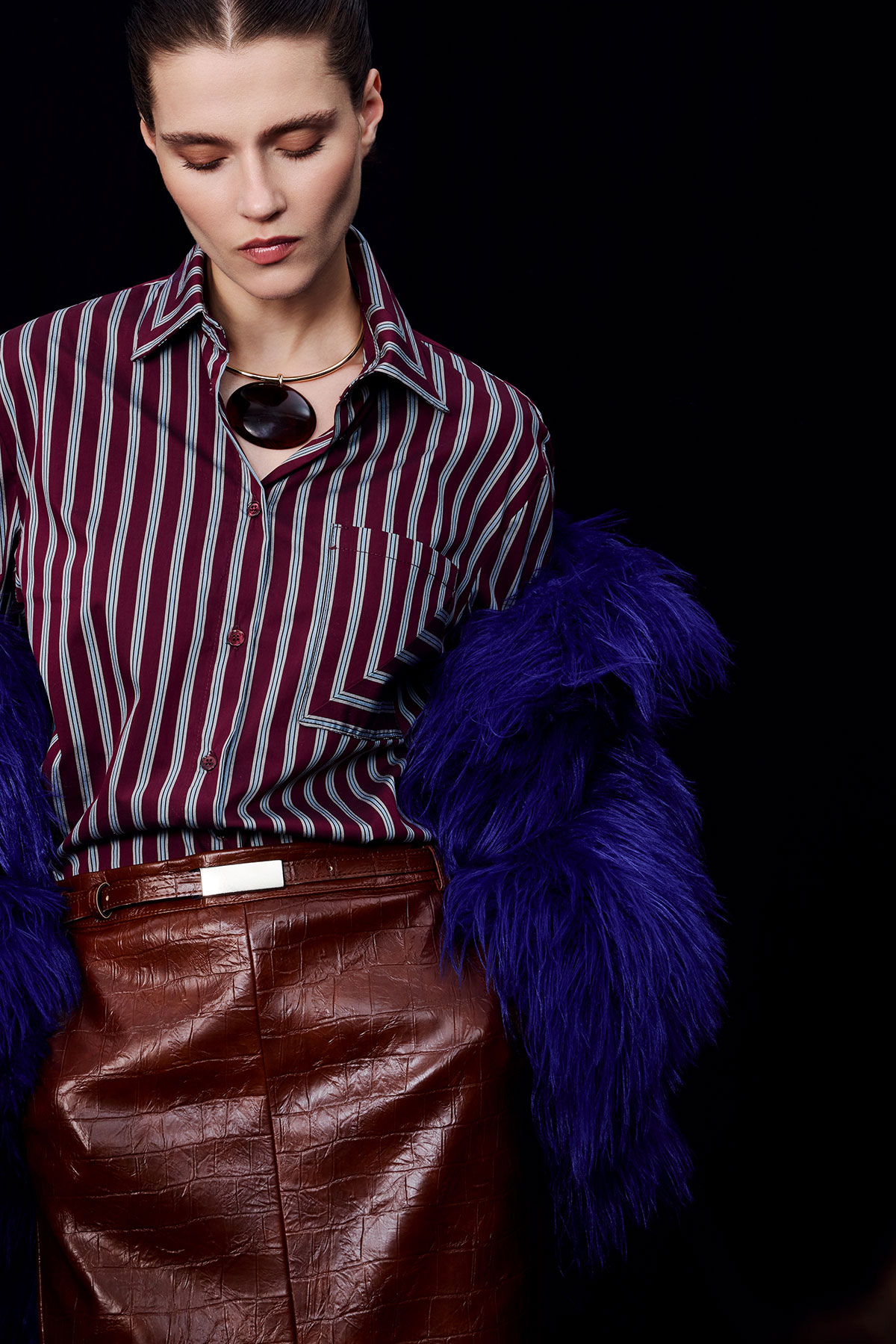 Woman in maroon striped shirt, brown leather skirt and purple faux-fur coat draped over arms, wearing a round pendant