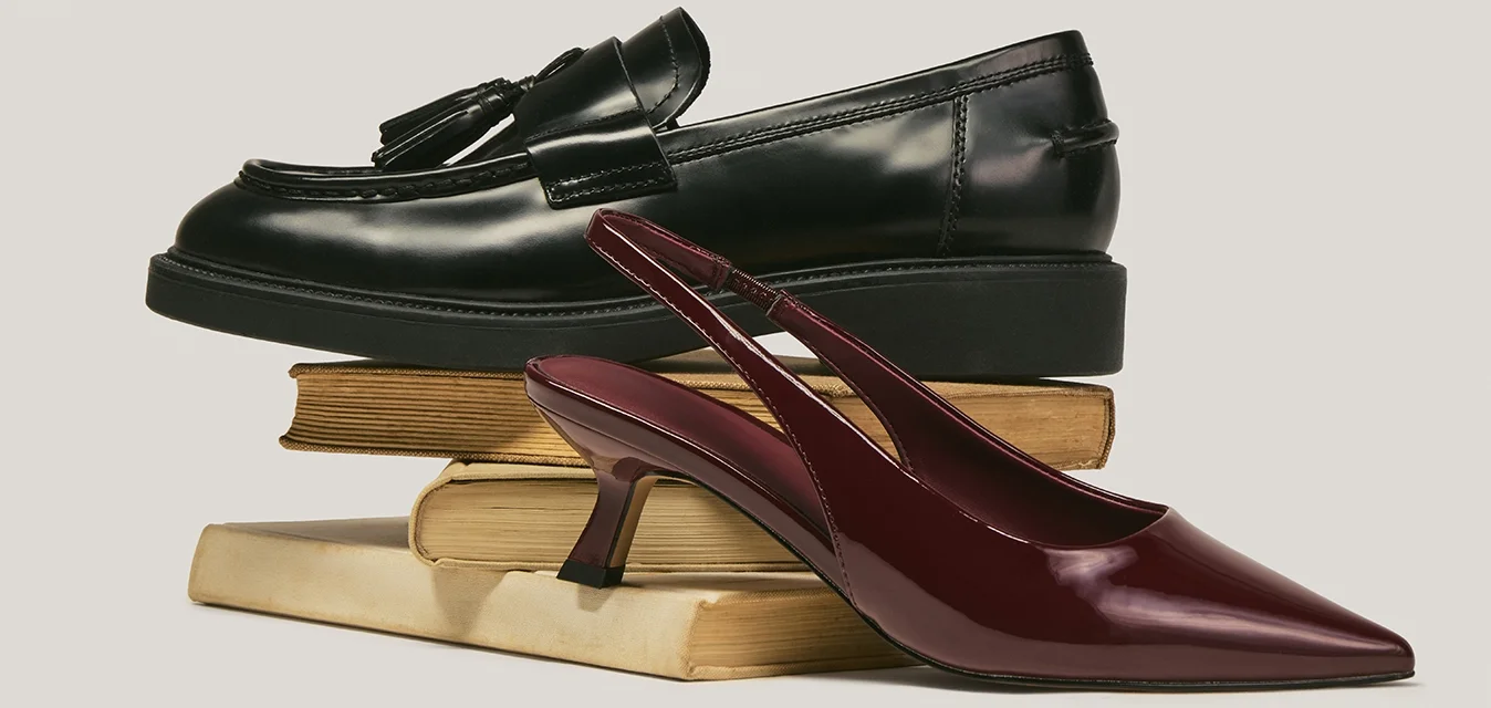 Mocassino in pelle nera e scarpa bassa con tacco basso appuntito color bordeaux impilati su due libri chiusi con copertina beige su sfondo neutro.