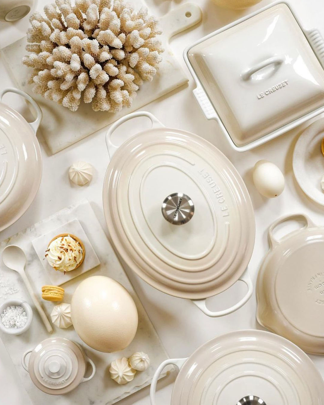 Overhead flat-lay of cream Le Creuset cookware and ceramic dishes with decorative coral, eggs and pastries on a white tabletop