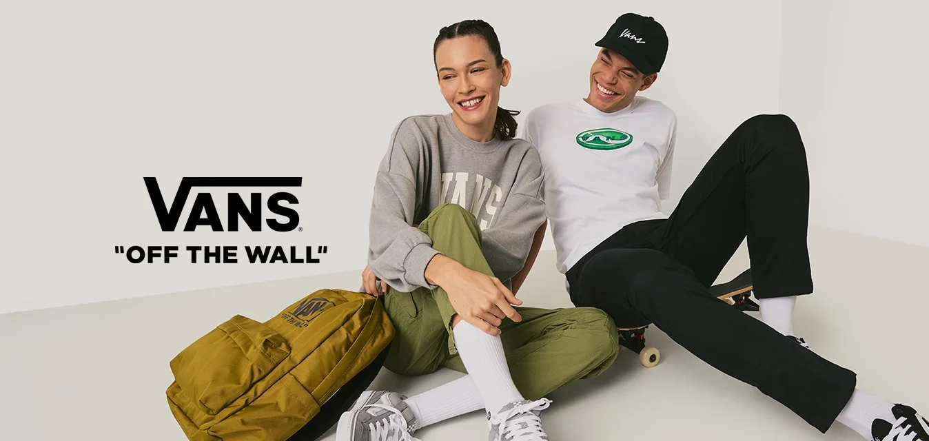 Due giovani seduti per terra in abiti casual, uno con uno skateboard e uno zaino giallo, sorridenti e vicini tra loro.