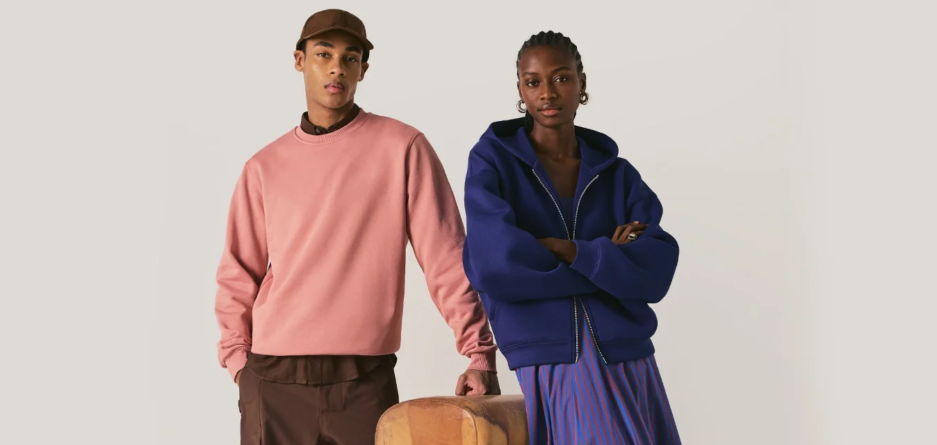 Uomo con cappello marrone, felpa rosa e pantaloni marroni in piedi accanto a una donna con felpa con cappuccio blu e gonna a righe vicino a un cavallo a pomolo di legno.