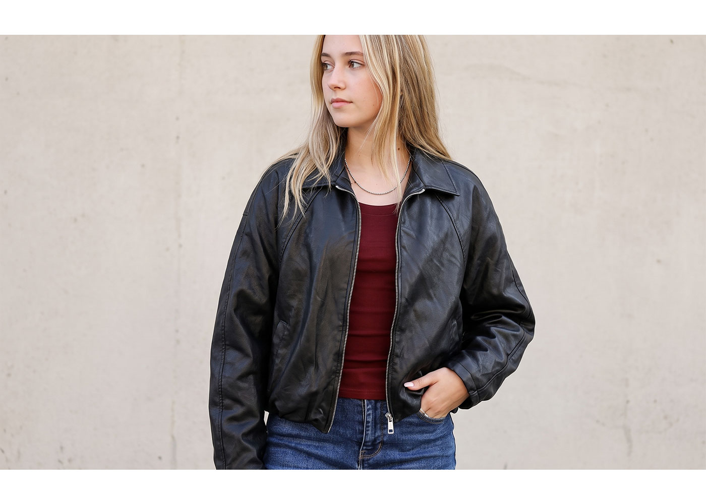 Young woman wearing a black leather jacket, burgundy top and blue jeans standing against a pale concrete wall with one hand in her pocket