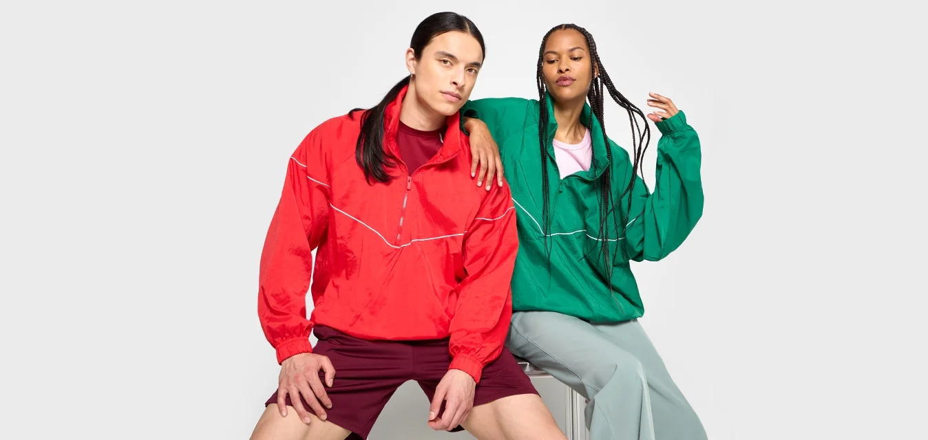 Due persone siedono su una sedia, indossando un giubbotto a vento rosso con dettagli bianchi e una giacca verde oversize, abbinate a pantaloni corti e pantaloni leggeri.
