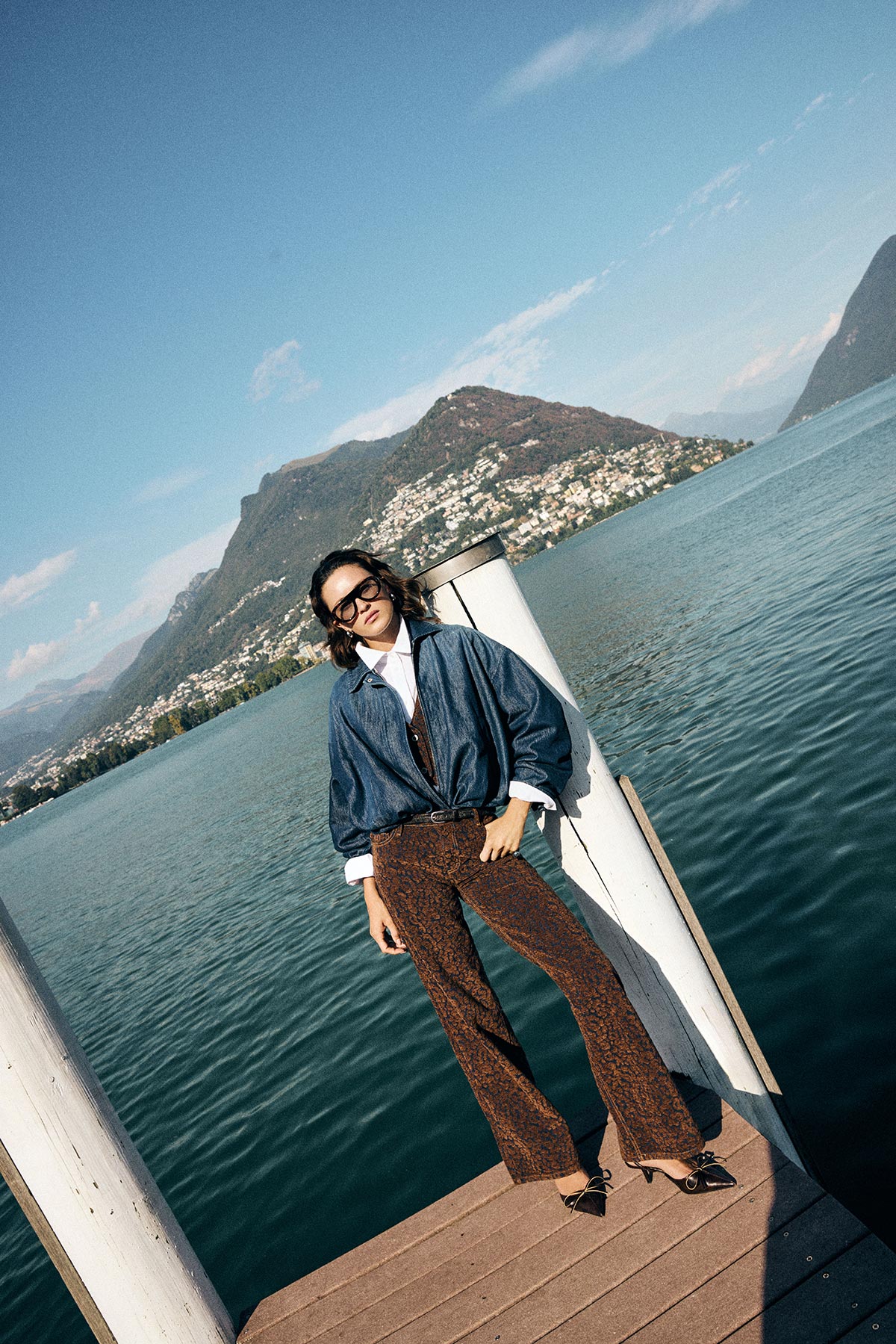 Woman in denim jacket, patterned brown flared trousers and sunglasses standing on a lakeside jetty with mountains and blue sky