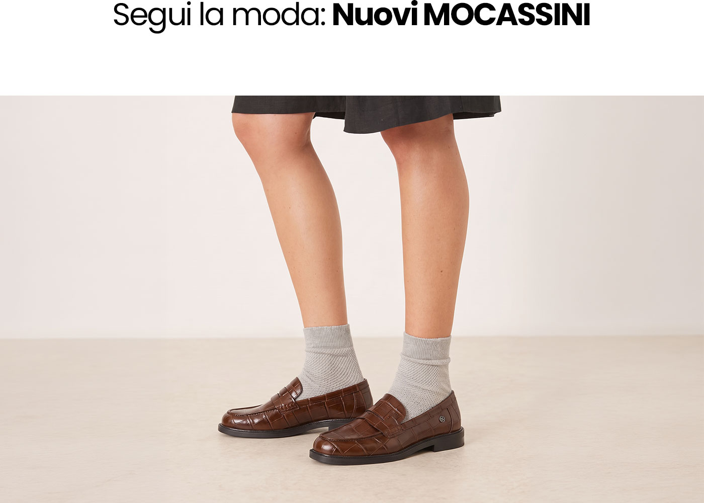 Brown croc-embossed leather loafers worn with grey socks and a black skirt on a neutral studio background