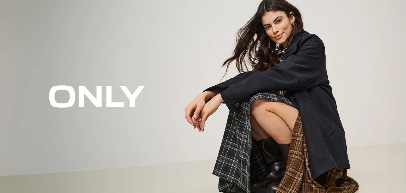 Donna con lunghi capelli scuri, indossa un cappotto nero e una gonna scozzese bicolore, accovacciata sul pavimento con stivali neri su uno sfondo chiaro e uniforme.