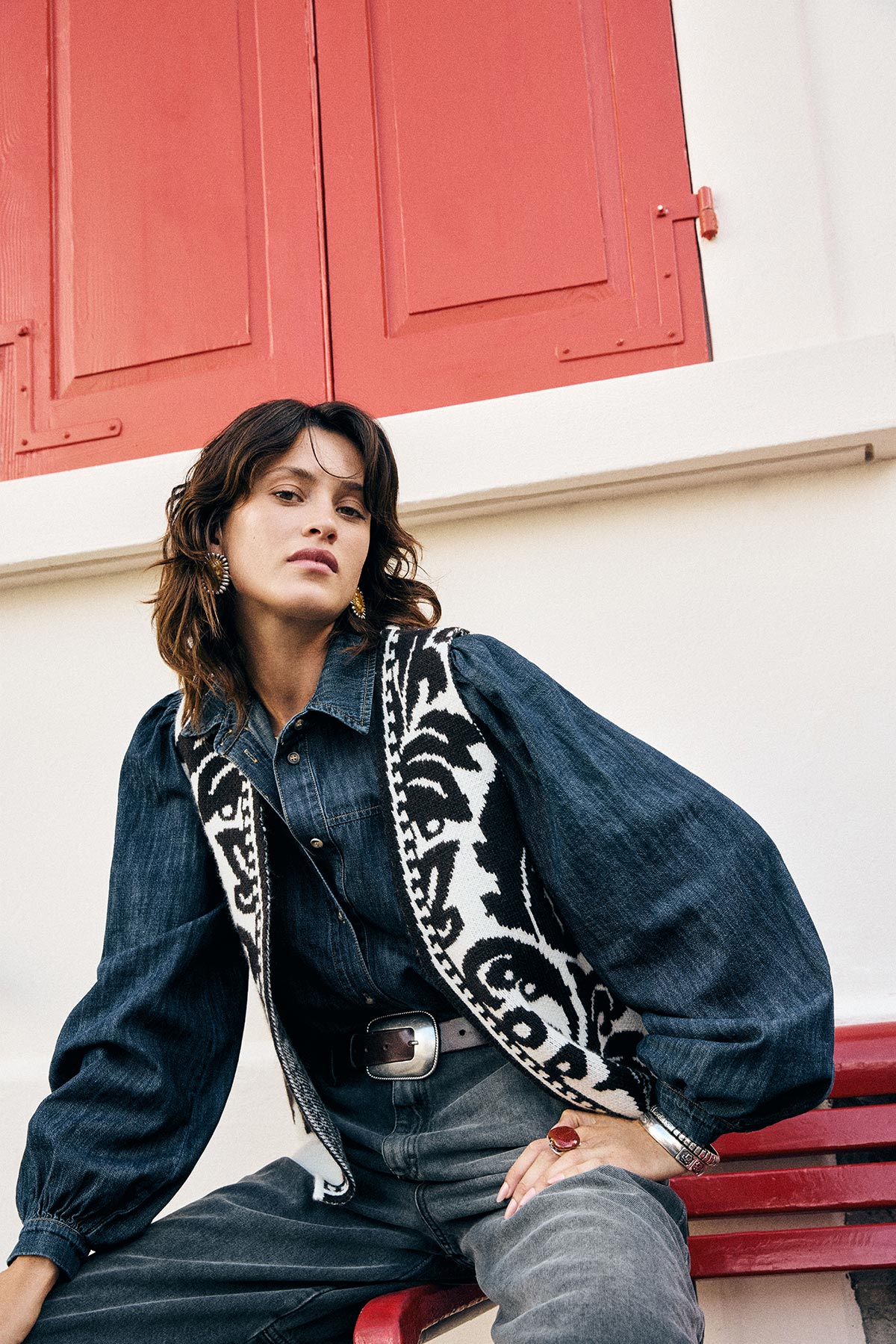 Woman wearing a denim shirt, patterned waistcoat and grey jeans seated on a red bench against a white wall with red shutters