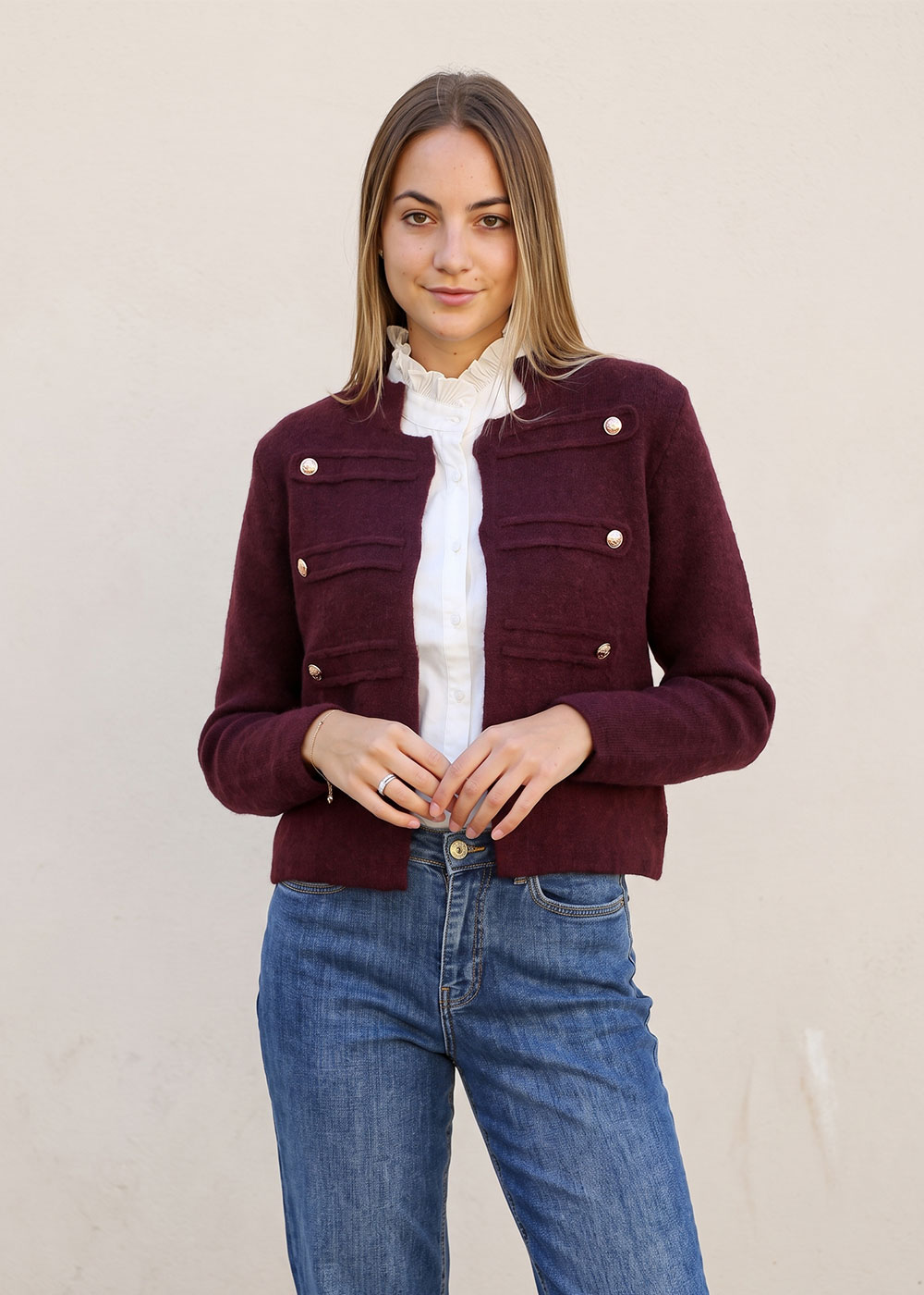 Young woman wearing burgundy military-style cropped jacket, white ruffled blouse and blue jeans against pale wall