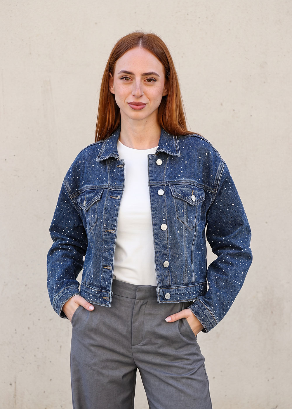 Young woman in speckled denim jacket, white top and grey trousers standing against a neutral wall with hands in pockets