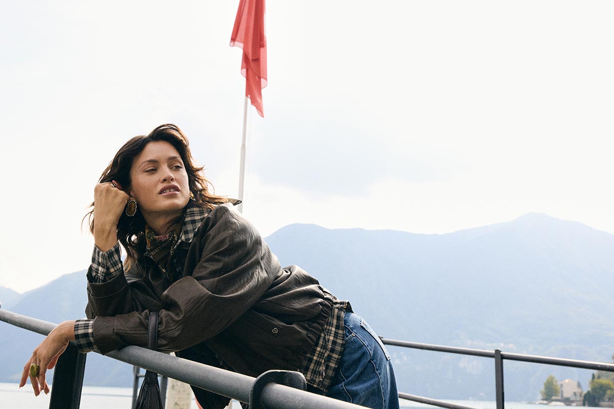 Woman in a brown leather jacket leaning on a railing by a lakeside with mountains in the background and a red flag