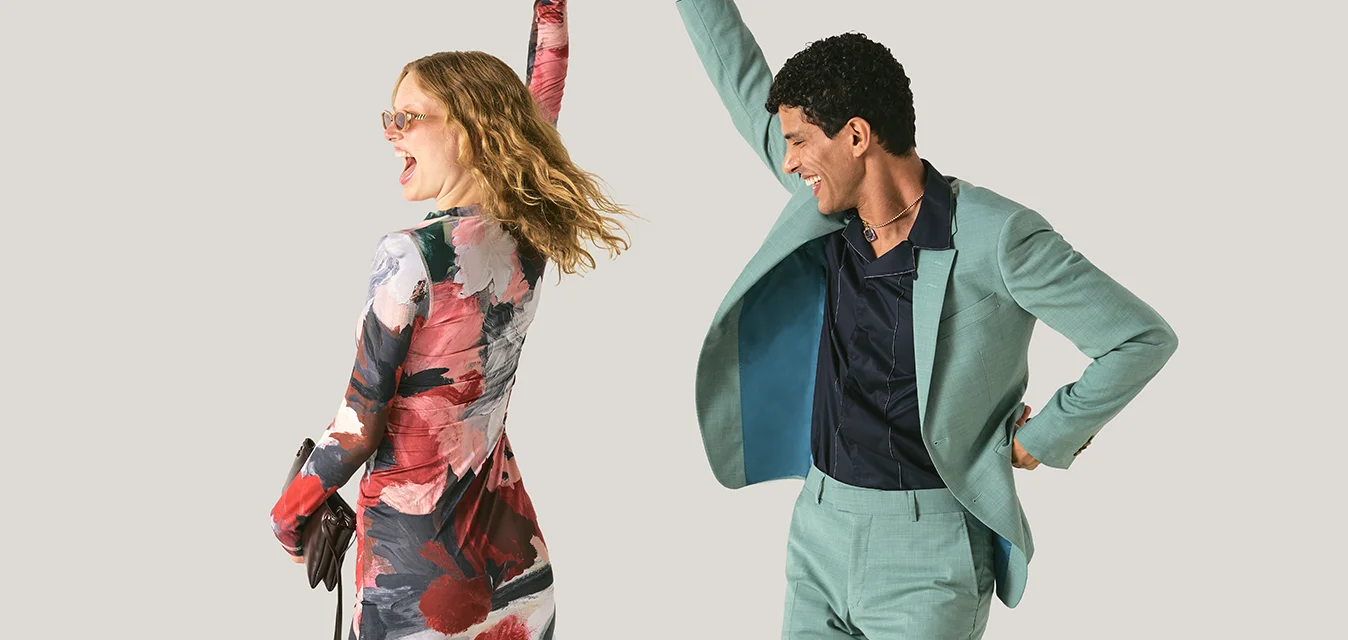 Un uomo in abito verde fa girare una donna con un vestito floreale colorato mentre ballano contro uno sfondo chiaro e semplice.
