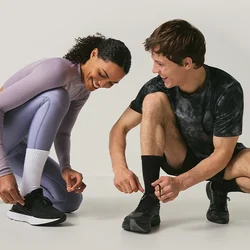 Due persone con abbigliamento sportivo sono accovacciate a legarsi le scarpe da corsa su uno sfondo semplice di colore chiaro.