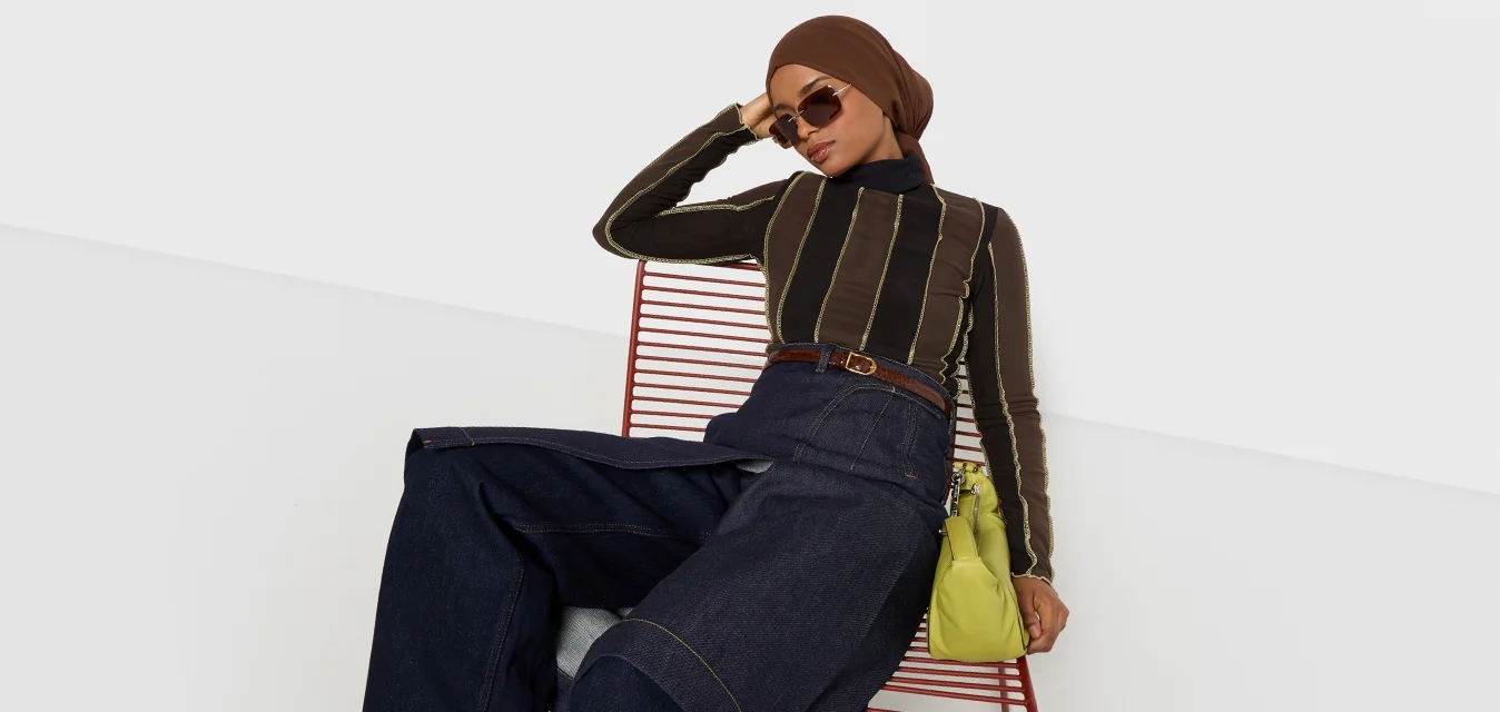 Top a maniche lunghe a righe marroni, pantaloni in denim a gamba larga a vita alta, borsa verde e occhiali da sole, seduta su una sedia di metallo rossa su sfondo bianco.