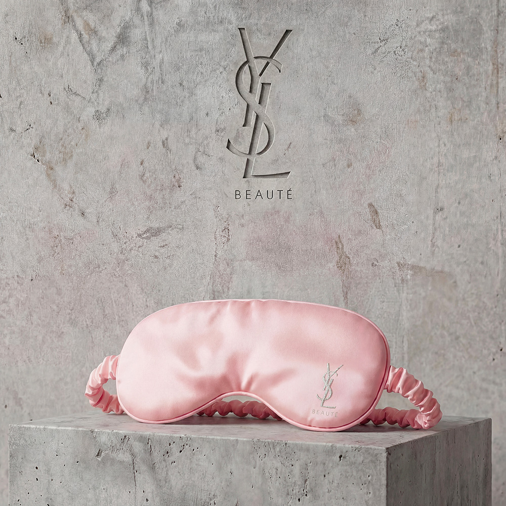 Una lussuosa mascherina da notte in raso rosa chiaro ricamata con il logo YSL Beaut&eacute;, appoggiata su un blocco di cemento.