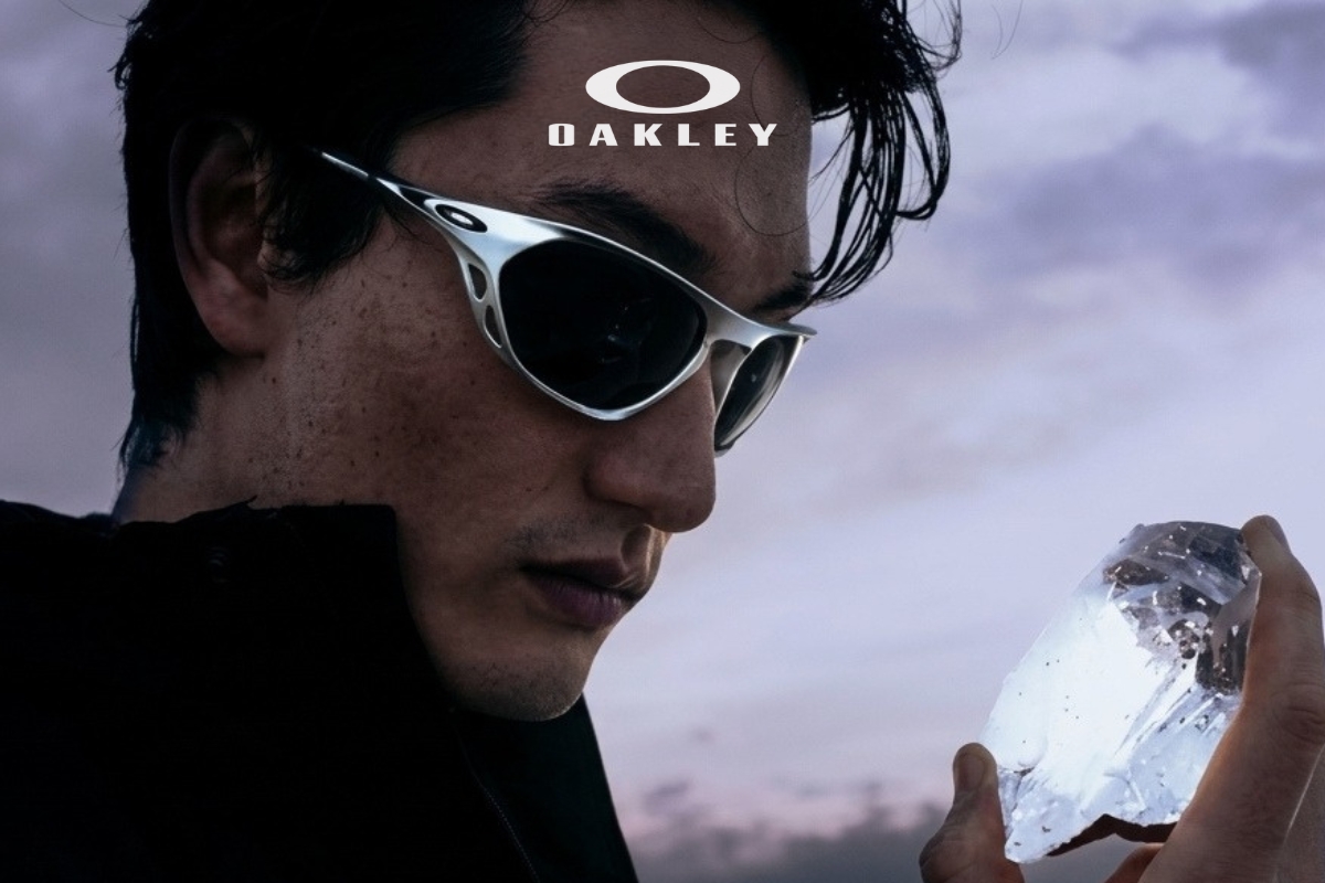 Close-up of a man wearing Oakley sunglasses holding a large clear crystal against a dusky sky, lifestyle product shot