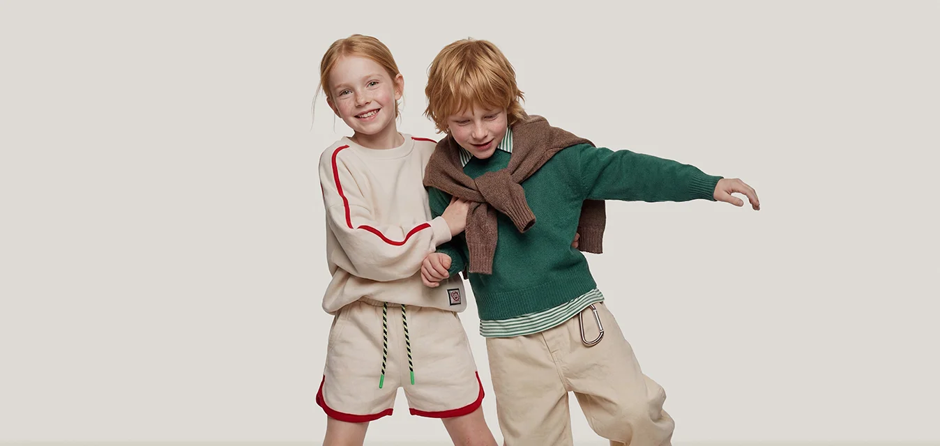 Due bambini che si tengono per mano e giocano, uno indossa pantaloncini beige e maglione, l'altro pantaloni beige con maglione verde e scarpe marroni.