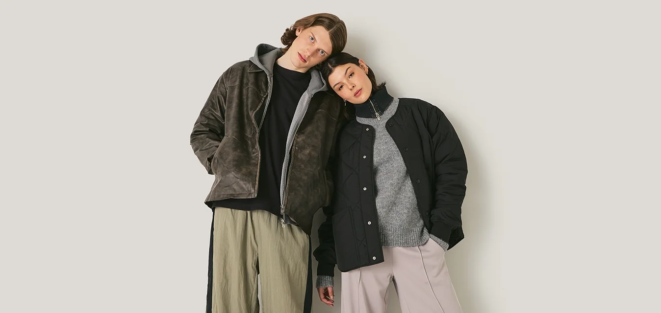 Due modelli si appoggiano l'uno all'altro, indossando una giacca marrone con texture, una giacca nera trapuntata, un maglione grigio e pantaloni palazzo in rosa chiaro e oliva.