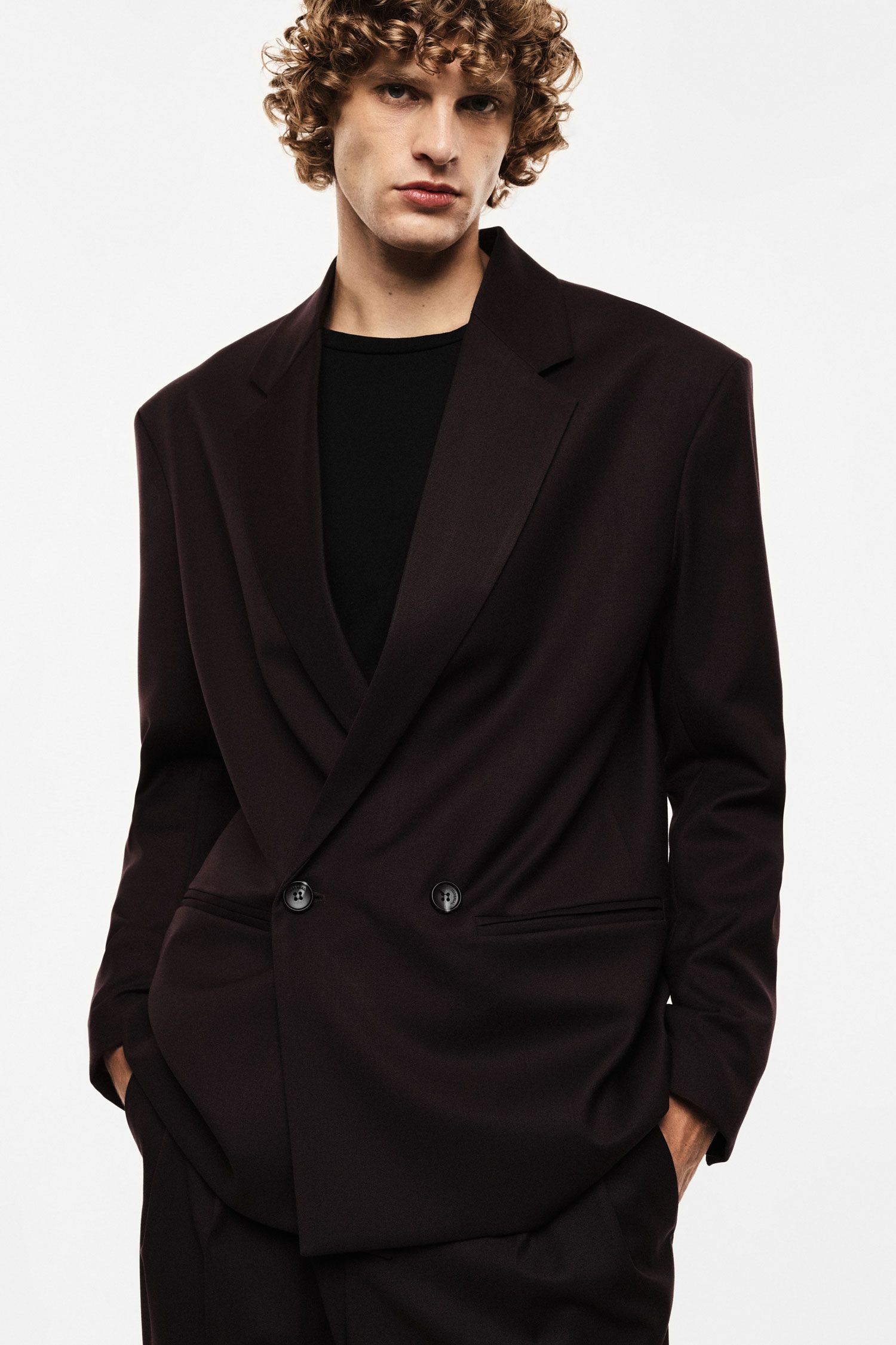 Young man with curly hair wearing a dark brown double-breasted blazer and black top, hands in pockets against plain white background