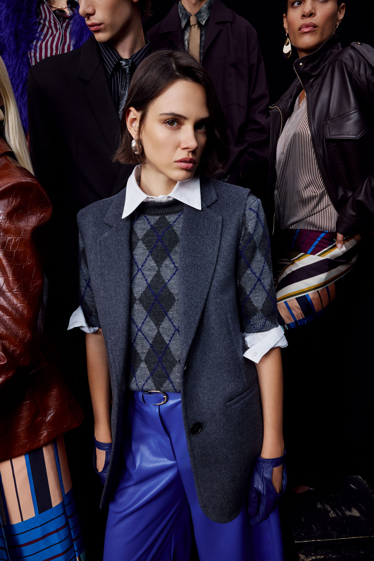 Fashion models grouped in studio, focus on woman in grey argyle sweater, sleeveless blazer, white shirt and blue leather trousers
