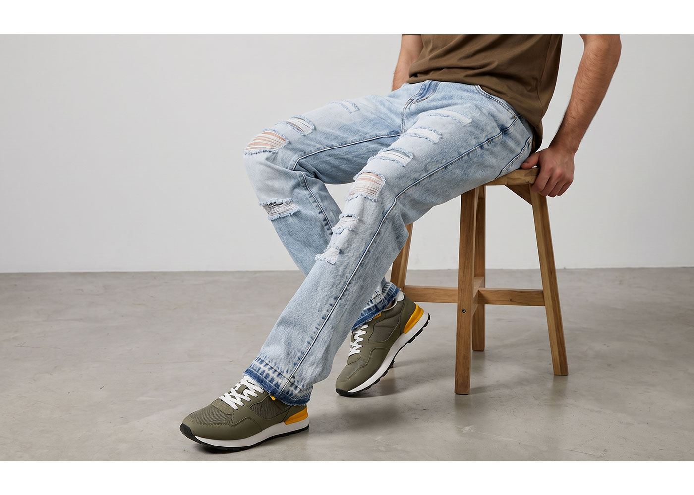 Person seated on wooden stool wearing ripped light-blue jeans and olive-green sneakers with yellow accents on a concrete floor