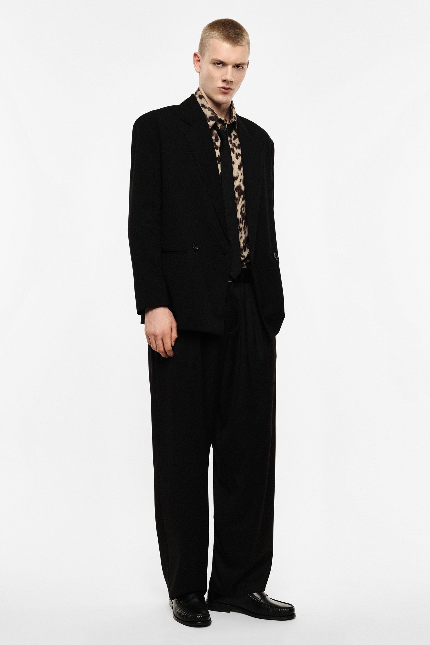 Male model wearing oversized black suit and leopard-print shirt, standing on plain white background