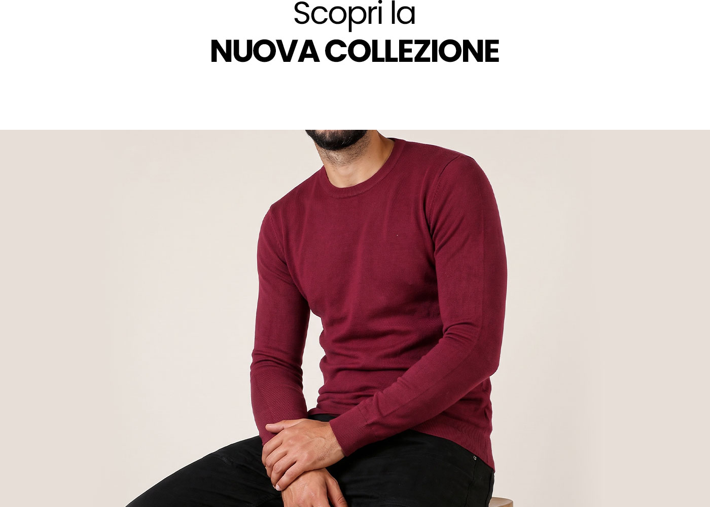 Seated male model wearing a burgundy crew-neck sweater and black trousers against a plain background, new collection