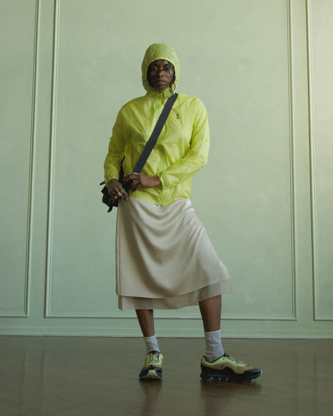Person wearing lime rain jacket with hood, cream mid-length skirt, white socks and yellow trainers, crossbody bag, standing indoors