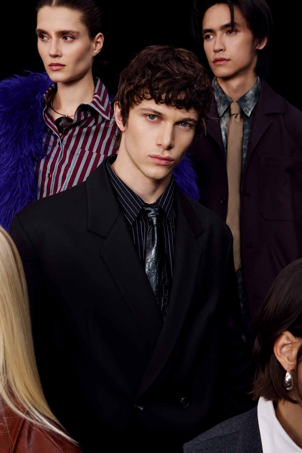 Three young fashion models in tailored suits and patterned shirts posing against a dark background, contemporary editorial style