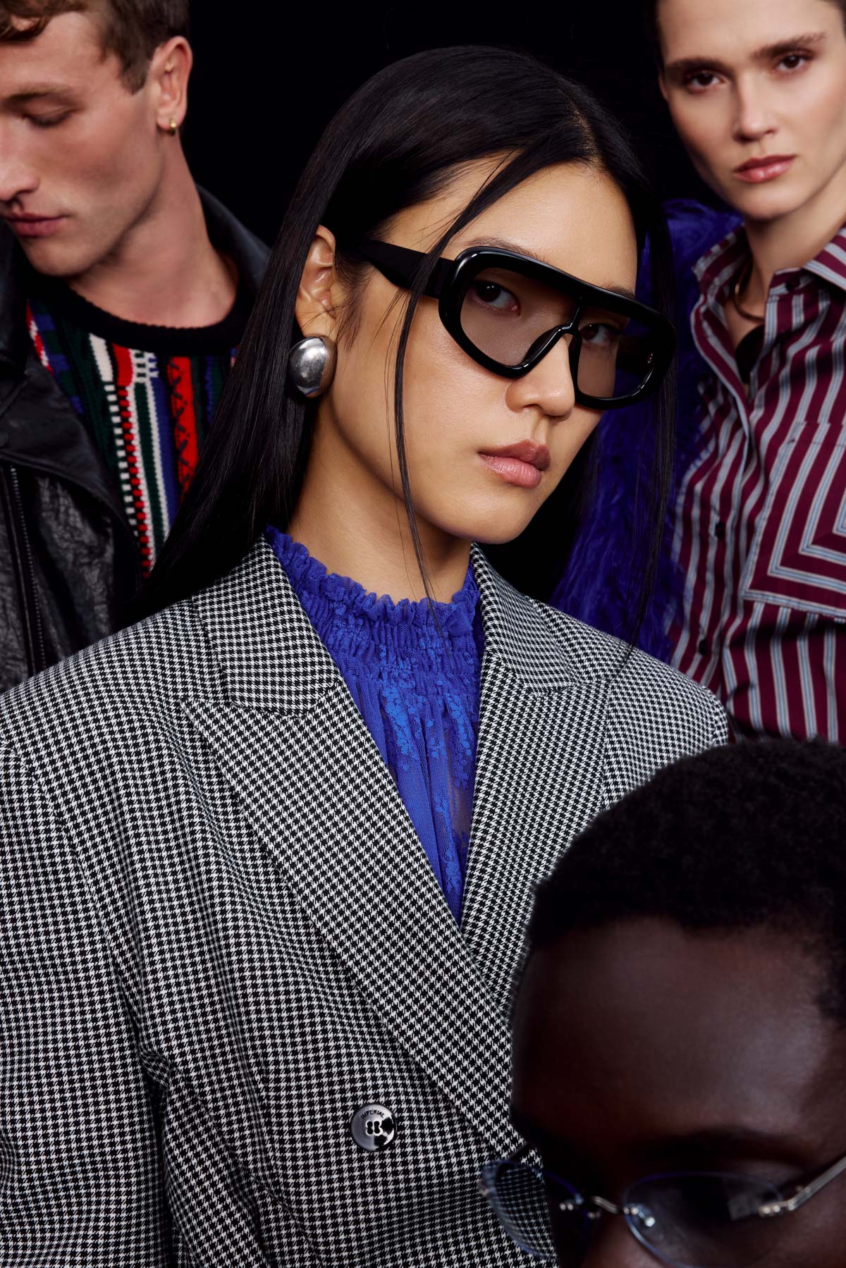 Group of fashion models; central woman with oversized black sunglasses, houndstooth blazer, blue lace top and silver earring