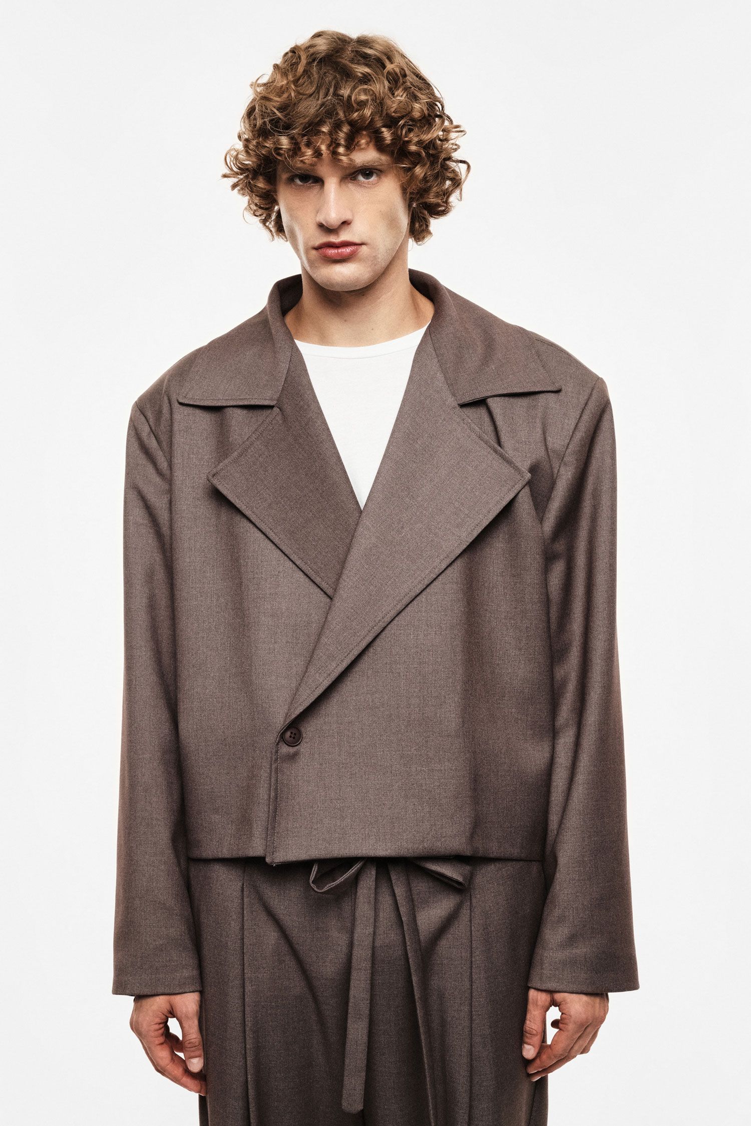 Young man with curly hair wearing a brown oversized cropped blazer and matching trousers on a white background