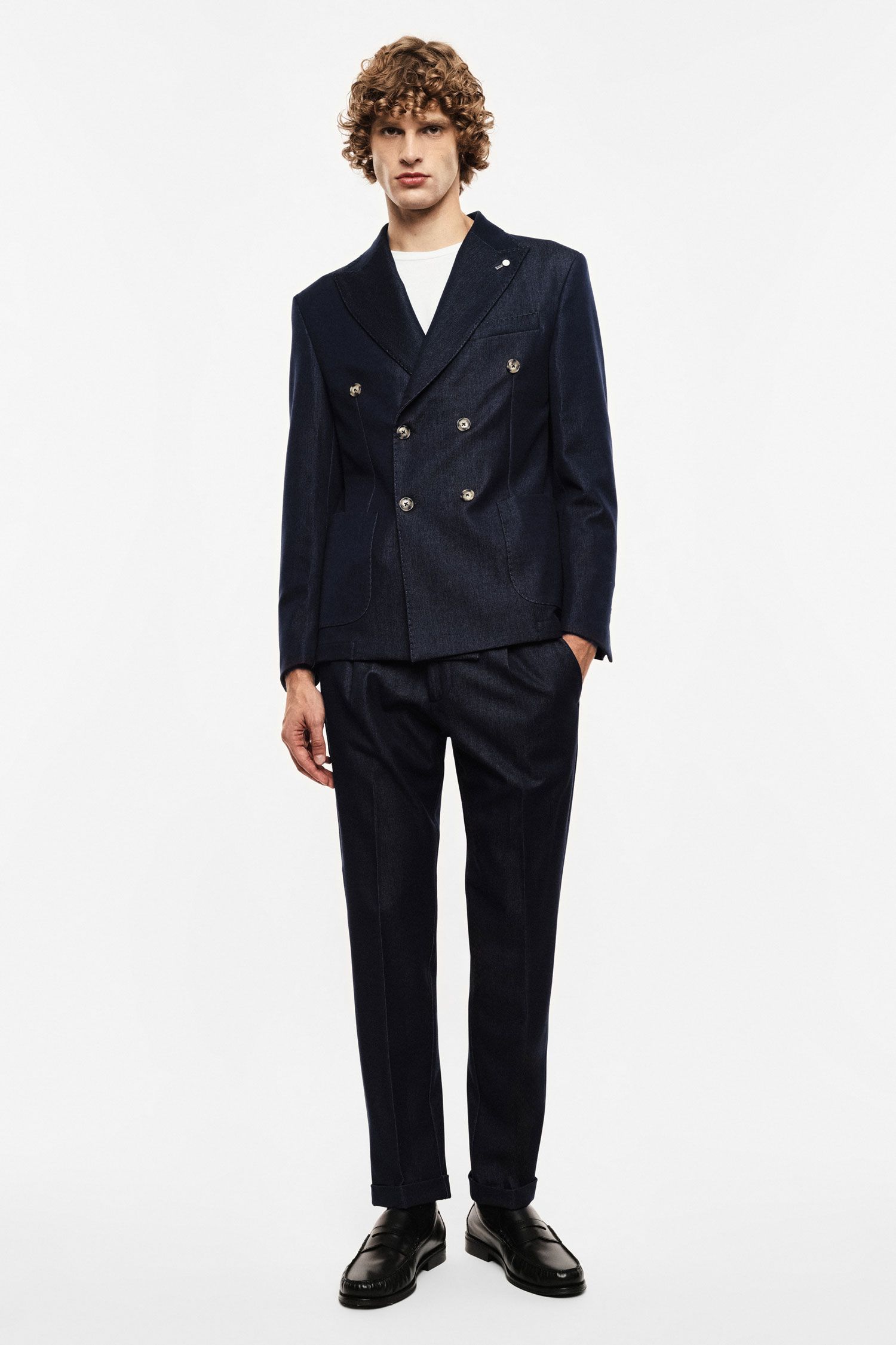 Male model in navy double-breasted suit, white T-shirt and black loafers, standing against plain white background