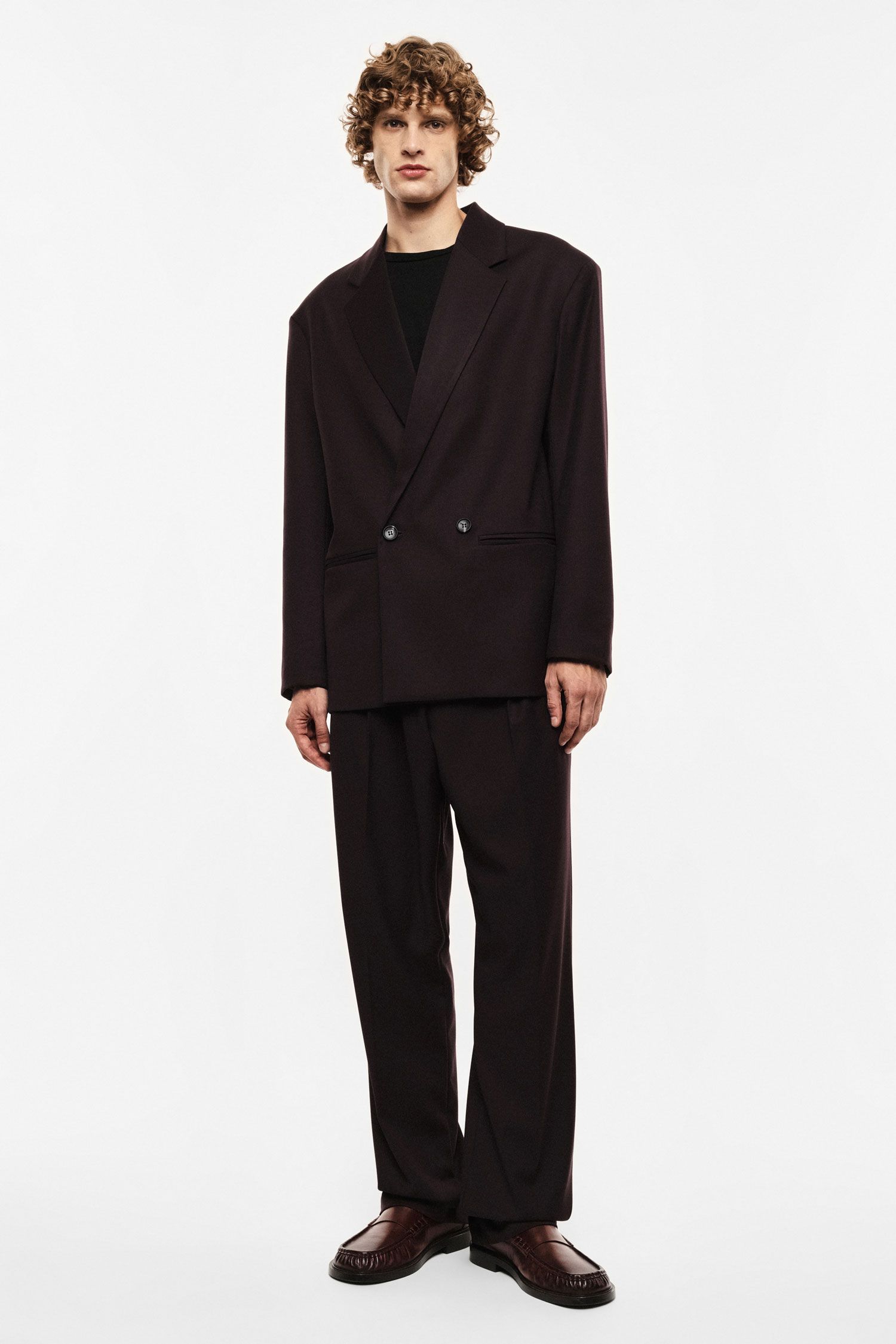 Male model with curly hair wearing oversized dark brown double-breasted suit, black top and brown loafers on white background
