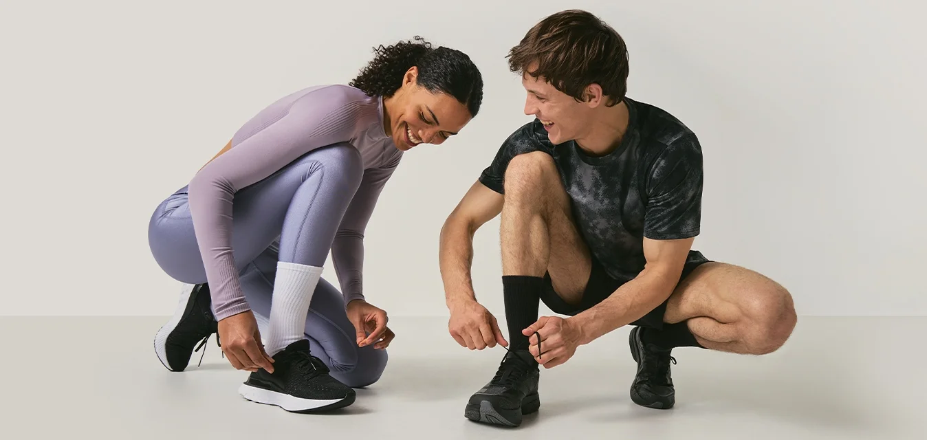 Due persone con abbigliamento sportivo sono accovacciate a legarsi le scarpe da corsa su uno sfondo semplice di colore chiaro.