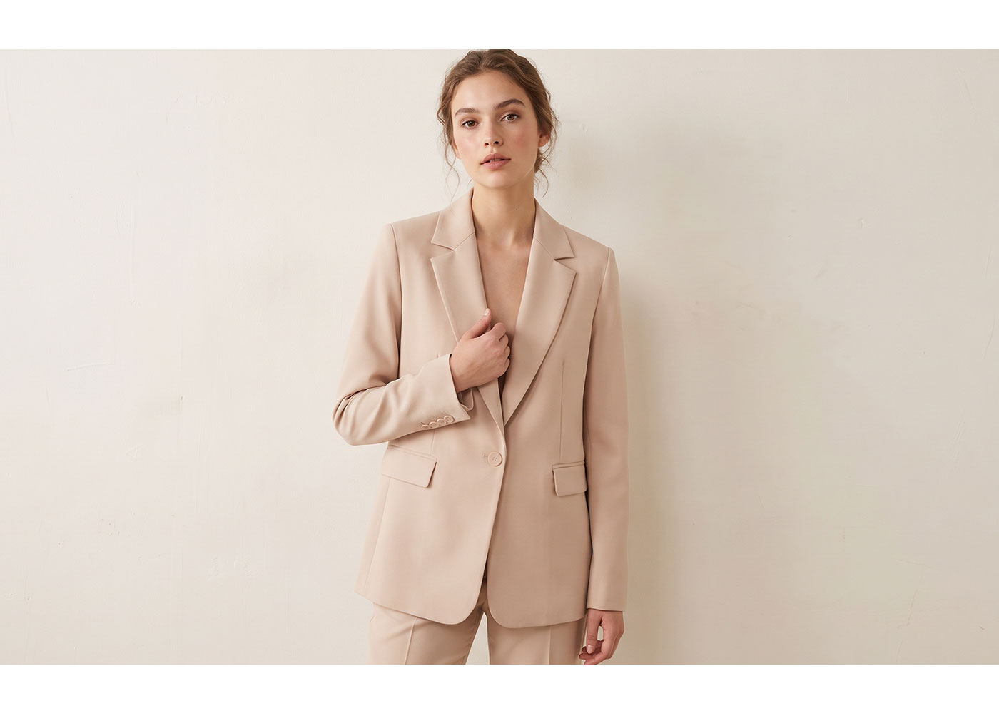 Young woman modelling a beige tailored blazer and matching trousers against a plain cream wall, minimalist fashion portrait
