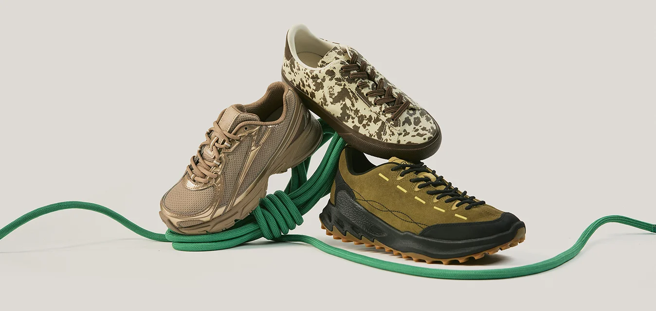 Tre sneaker eleganti in marrone, verde e beige con stampa mimetica poggiano in equilibrio su una corda verde contro uno sfondo semplice.