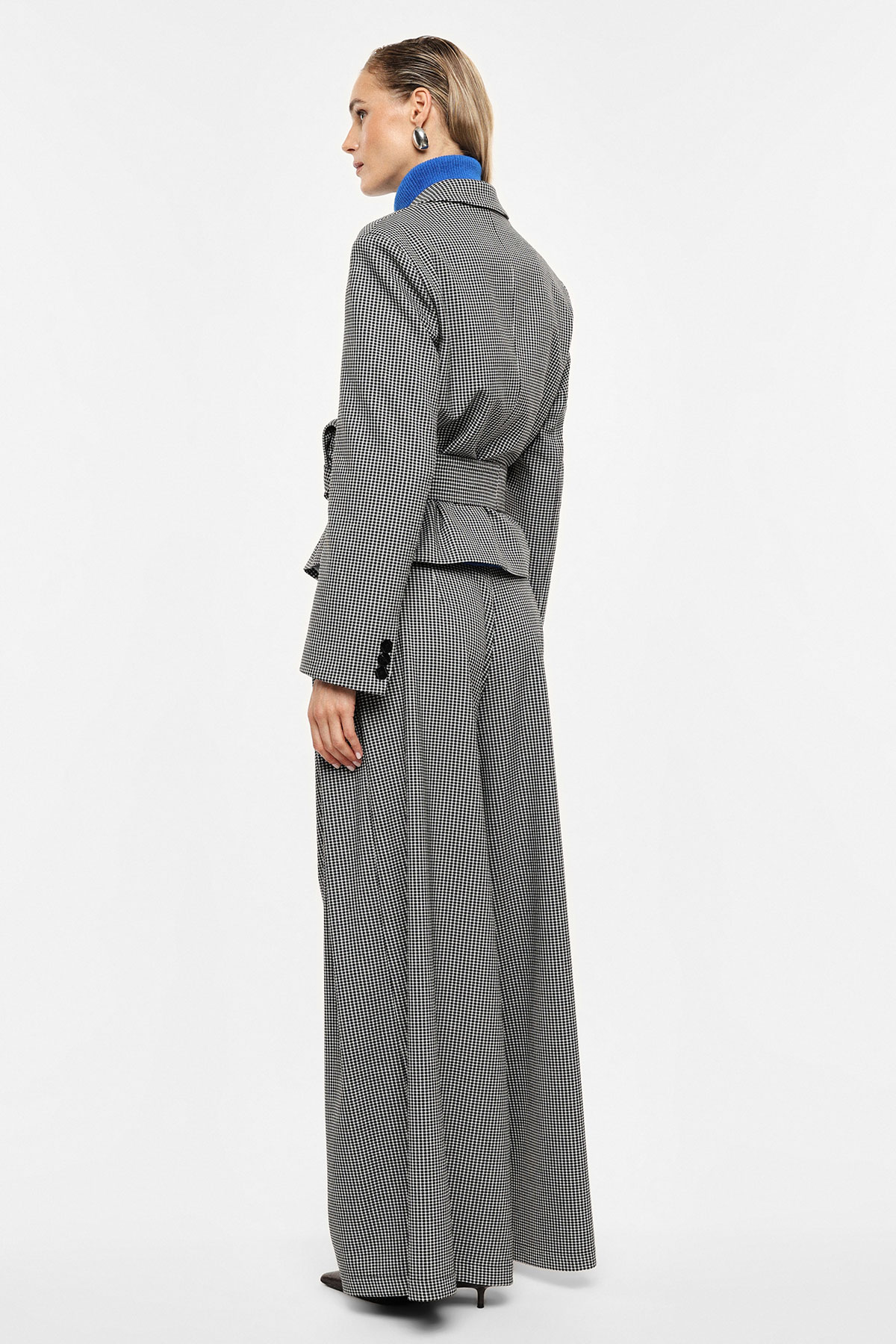 Rear three-quarter view of model wearing black-and-white houndstooth tailored blazer and matching wide-leg trousers with blue roll-neck