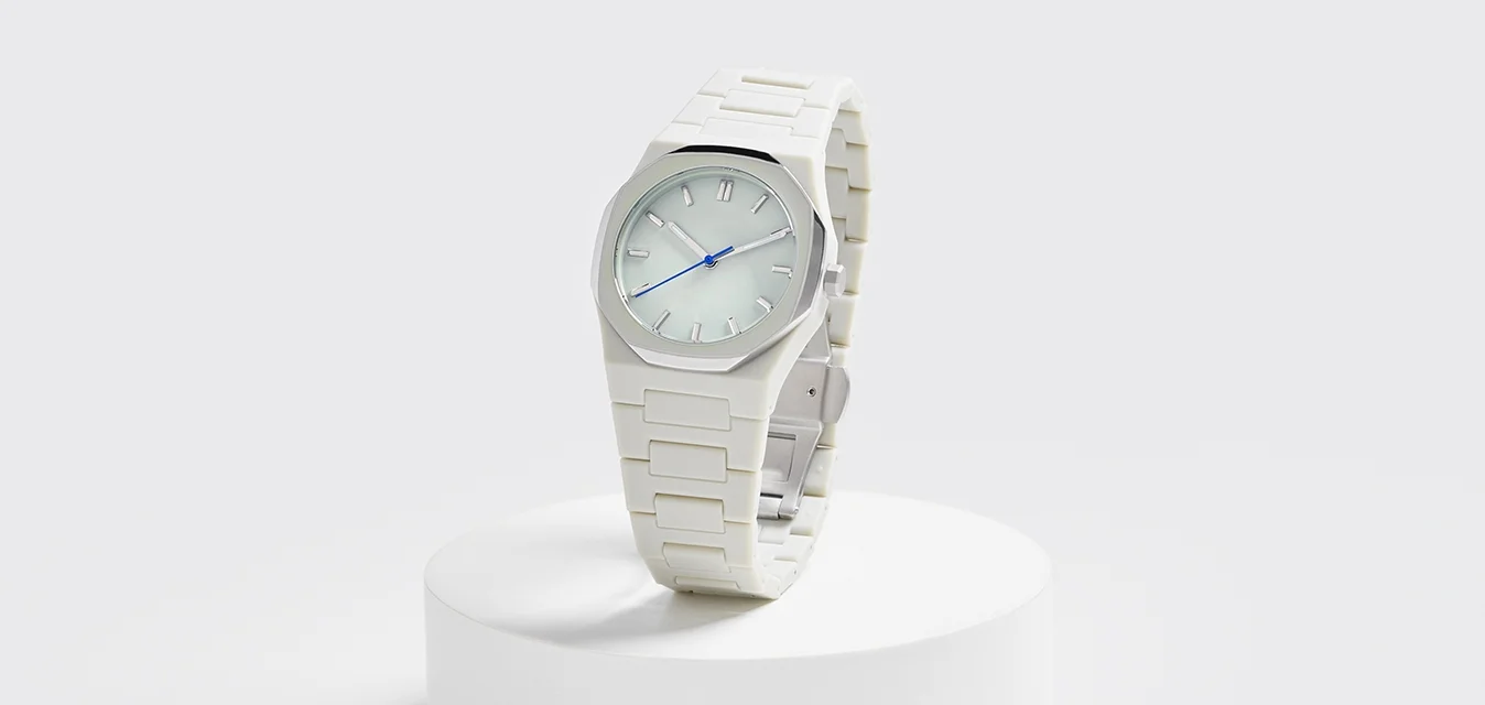 Orologio bianco in plastica con quadrante ottagonale liscio e lancette blu, esposto su un podio rotondo bianco contro uno sfondo chiaro.
