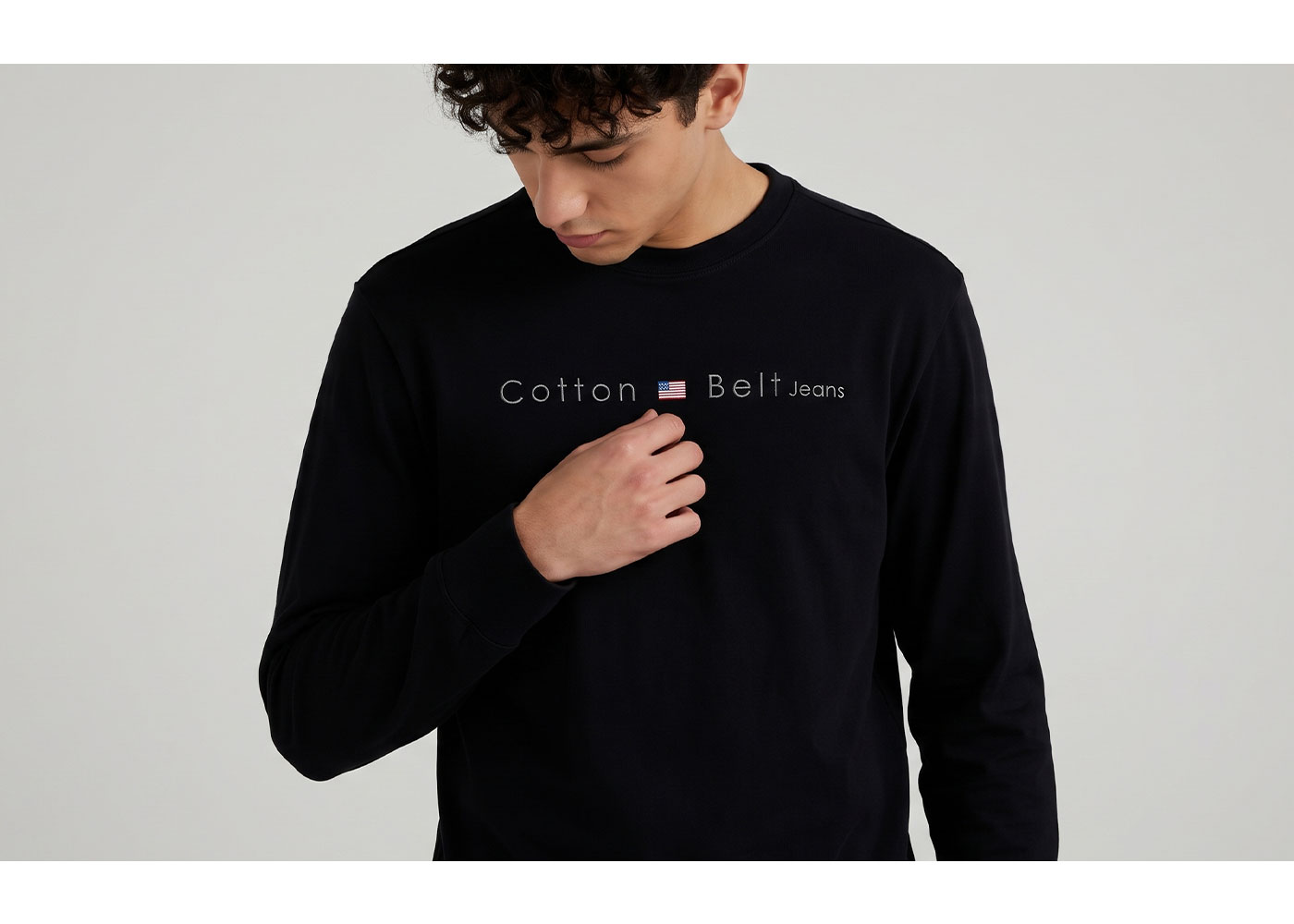 Young man wearing a black long-sleeve 'Cotton Belt Jeans' logo top with a small American flag, looking down and touching his chest
