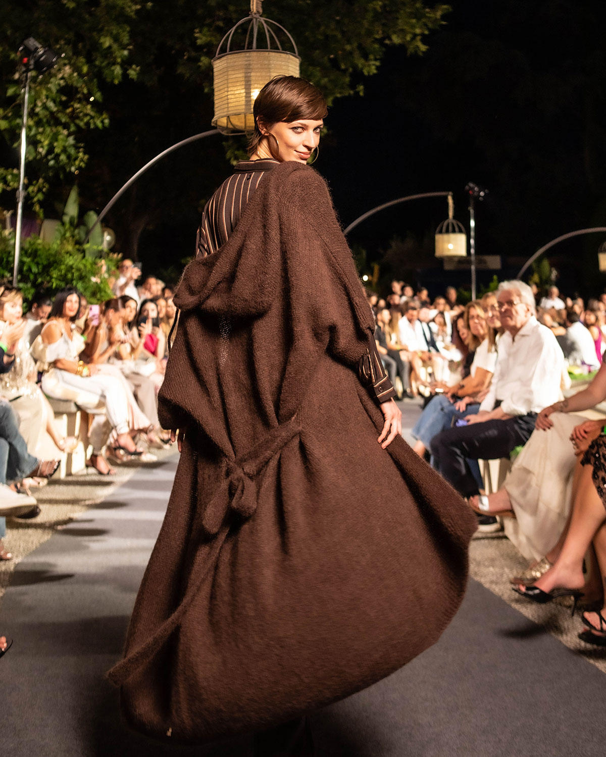 Model on outdoor evening runway, turning back in a flowing oversized brown coat at a fashion show with seated audience