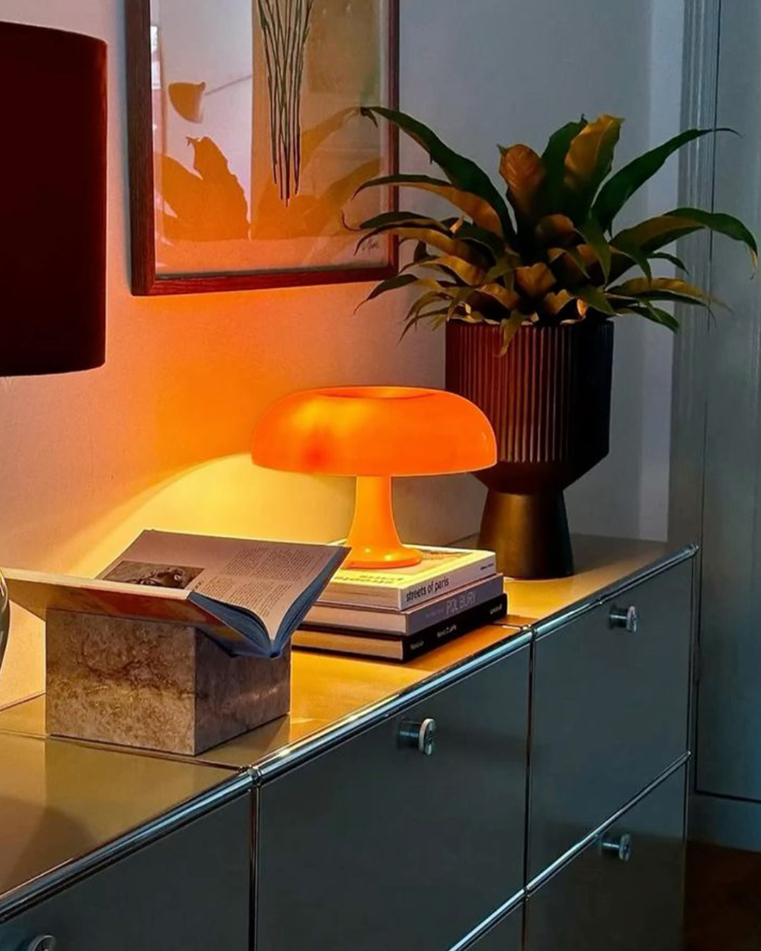 Orange mushroom table lamp glowing on a modern sideboard with stacked books, an open book, a potted plant and framed artwork