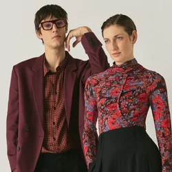 Due modelli si trovano di fronte a uno sfondo neutro; uno indossa un blazer bordeaux e una camicia a fantasia, l'altro una maglietta floreale rossa e pantaloni neri a vita alta.