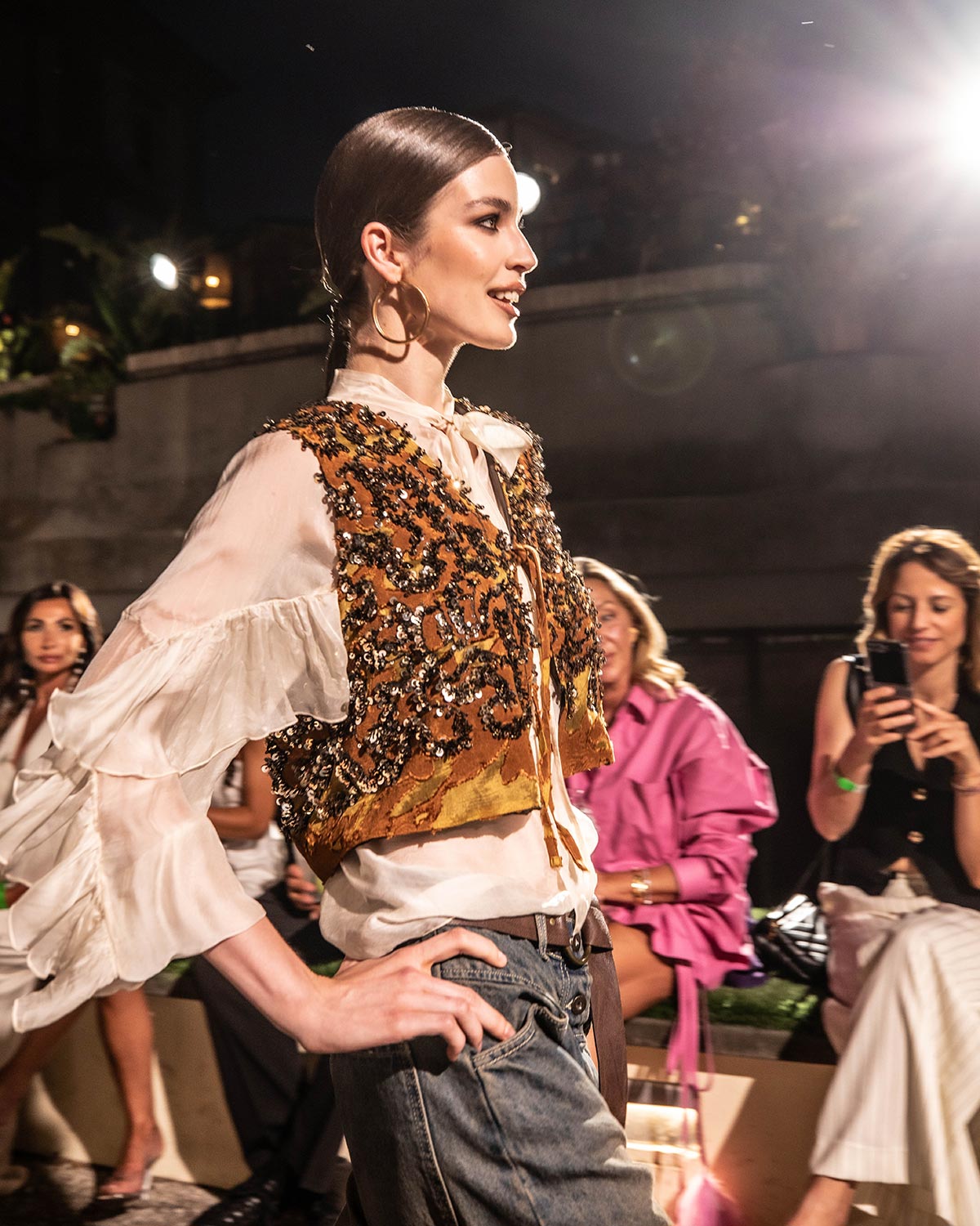 Runway model in an embellished gold vest, ruffled white blouse and jeans with hoop earrings walking past seated audience at night