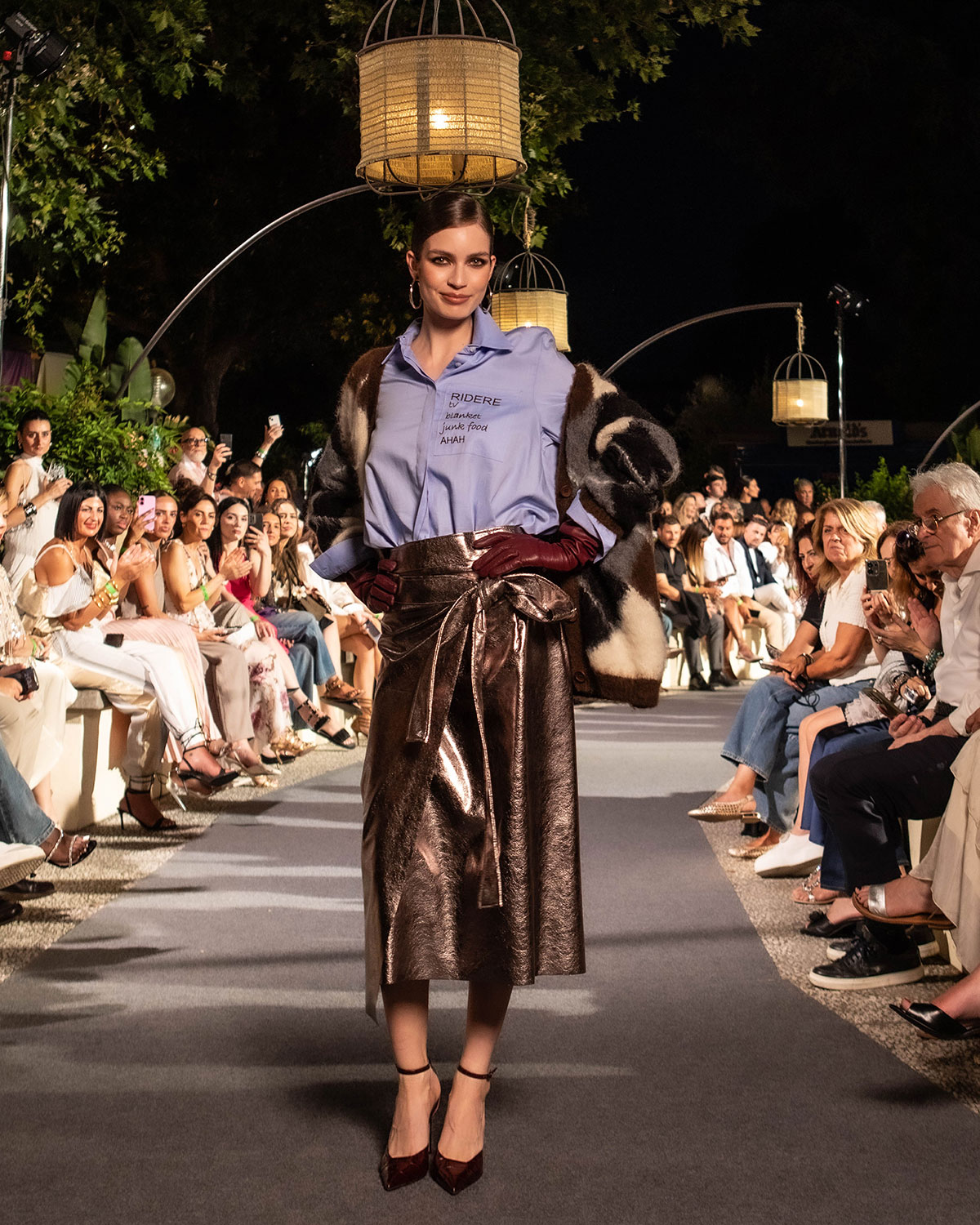 Model on outdoor runway wearing a blue shirt, patterned fur coat, metallic bronze wrap skirt and burgundy gloves as seated audience watches