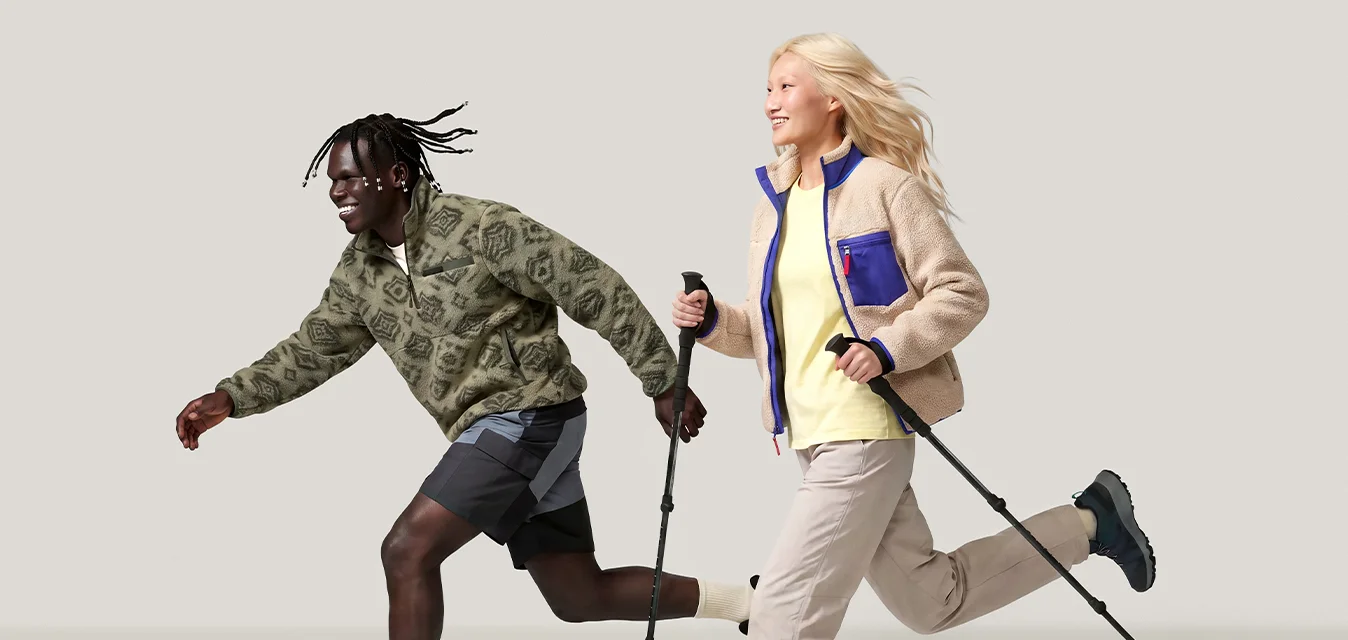 Due persone con abbigliamento casual da esterno che corrono gioiosamente; una usa i bastoncini da passeggio, l'altra corre accanto a loro con un'espressione sorridente.