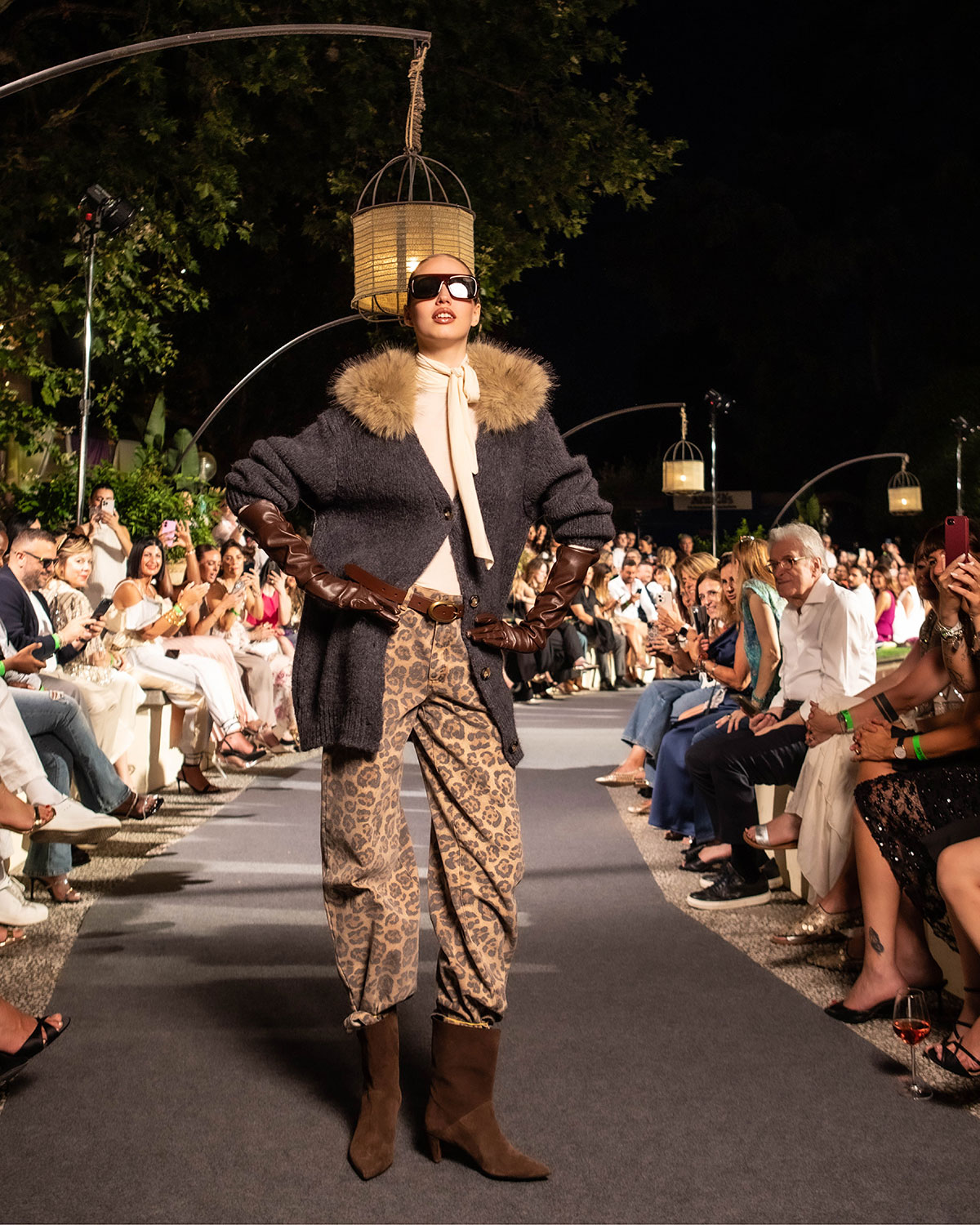 High-fashion runway model in fur-collared coat, long leather gloves and leopard-print trousers walking past seated audience