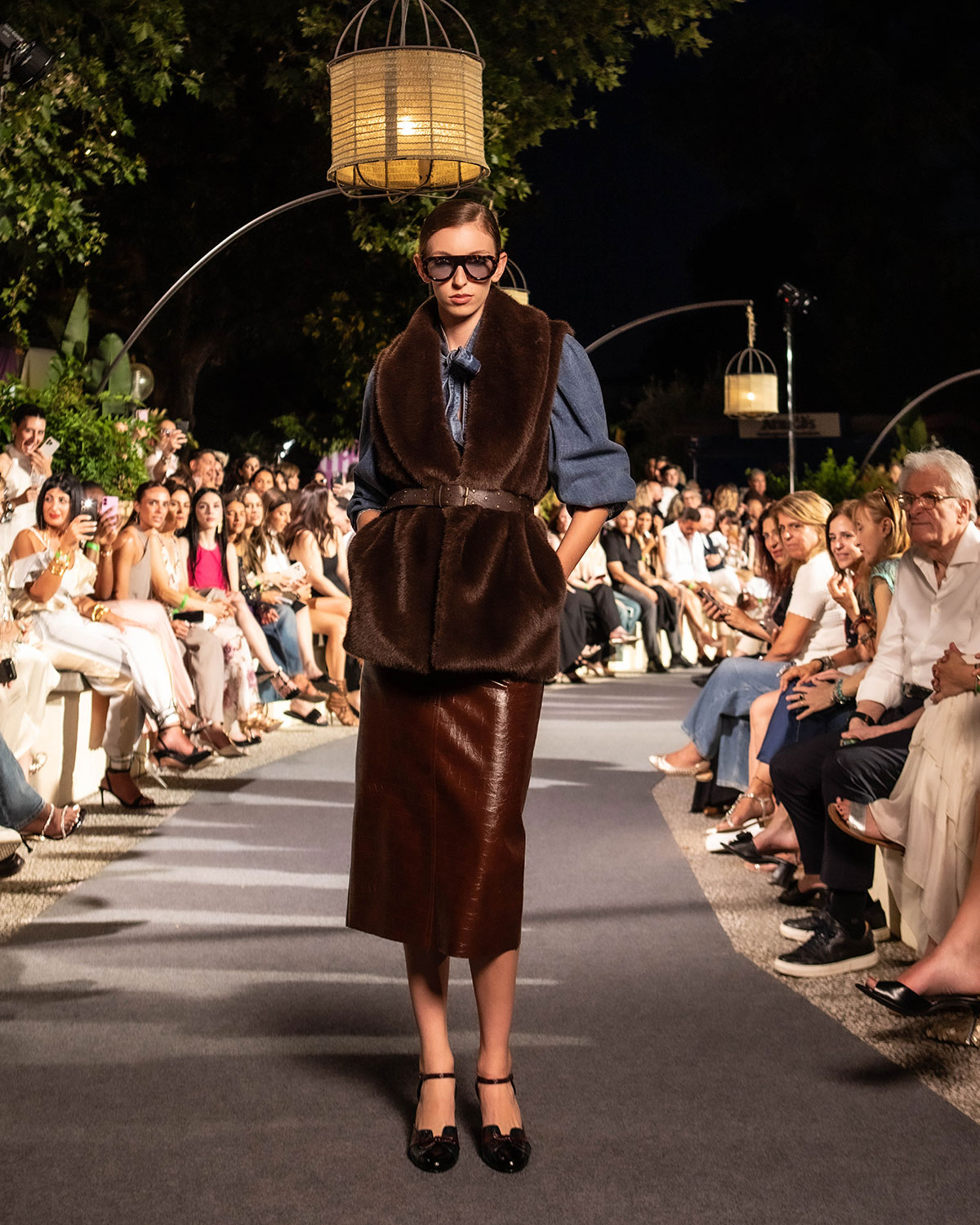 Model on outdoor evening runway wearing brown faux-fur vest, leather skirt and sunglasses, seated audience