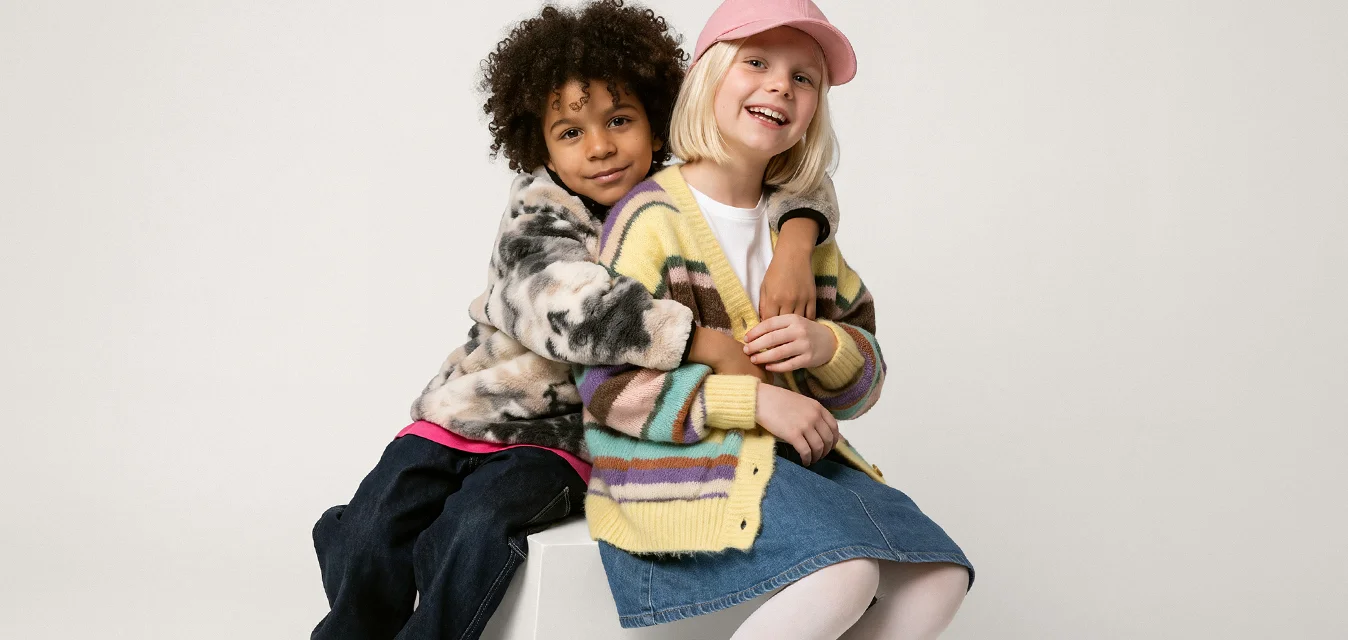 Due bambini seduti su un blocco bianco, uno abbraccia l'altro, entrambi sorridenti, vestiti con abiti e scarpe colorati e casual.