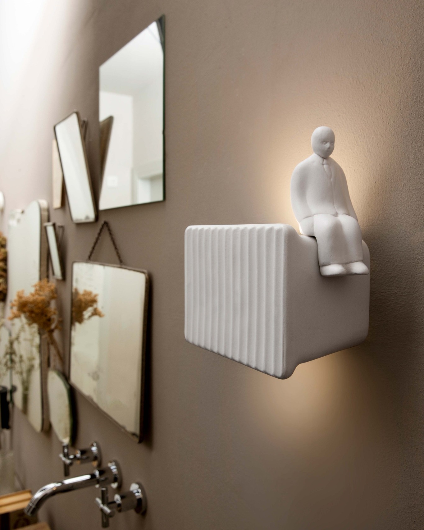 White ceramic wall light shaped as a seated figure on a ribbed block, mounted above bathroom taps and mirrors
