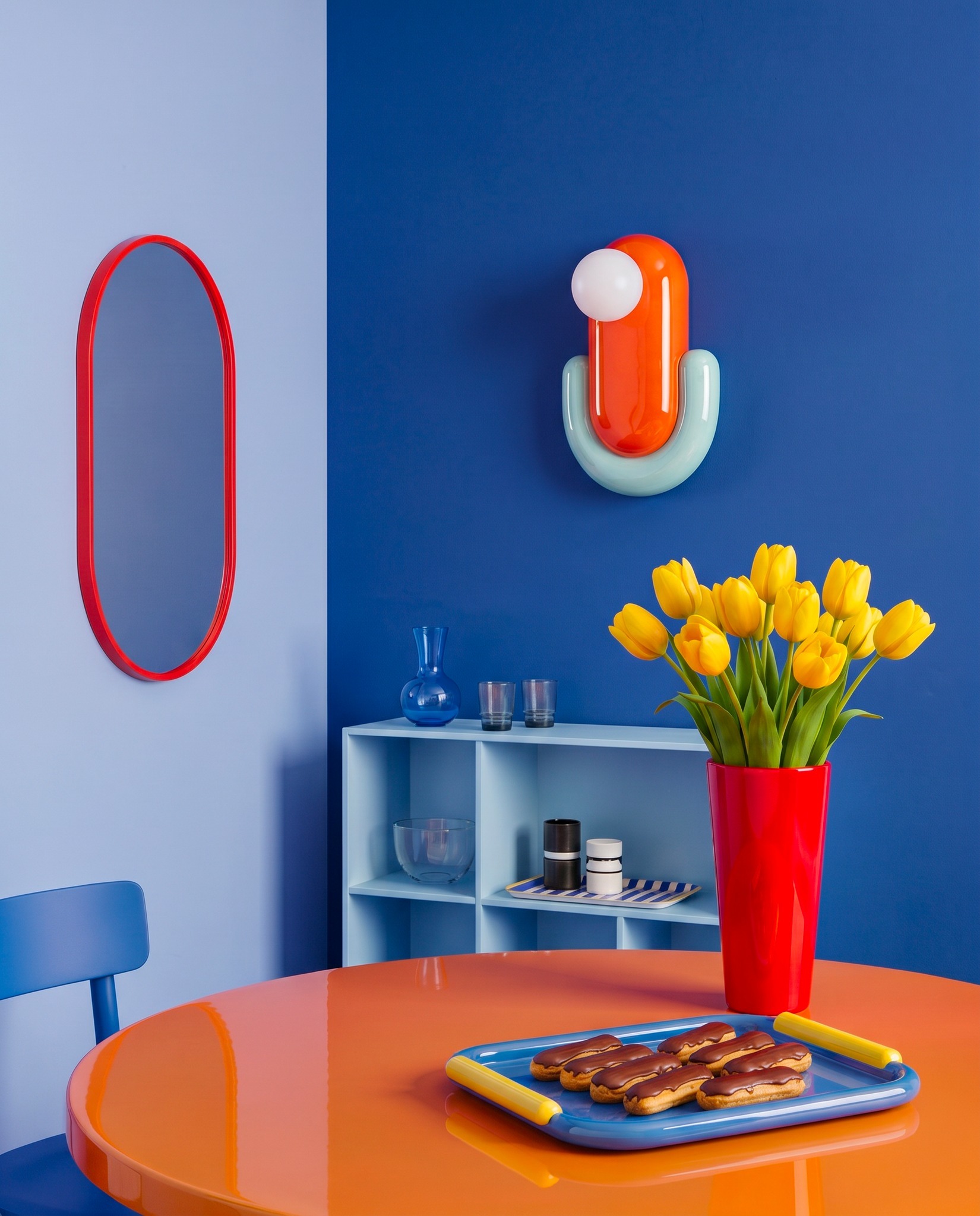 Bright modern dining scene with orange glossy table, blue tray of chocolate éclairs and red vase of yellow tulips against a blue wall